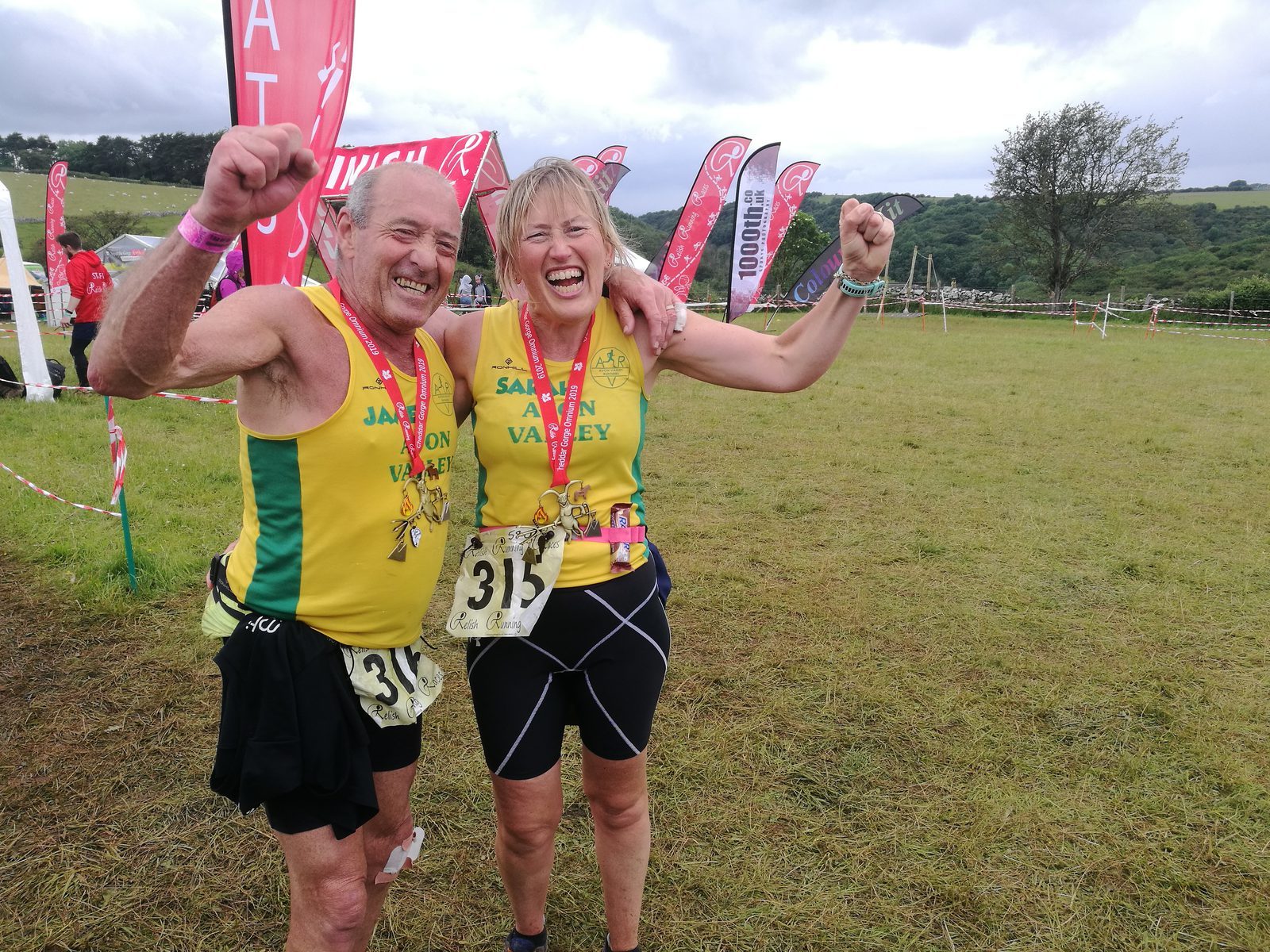 Two runners celebrate with their arms raised, both smiling and holding medals. They are wearing matching yellow and green athletic jerseys with black shorts. The woman has race number 315 and the man has number 316. Red flags and a grassy field are visible in the background.