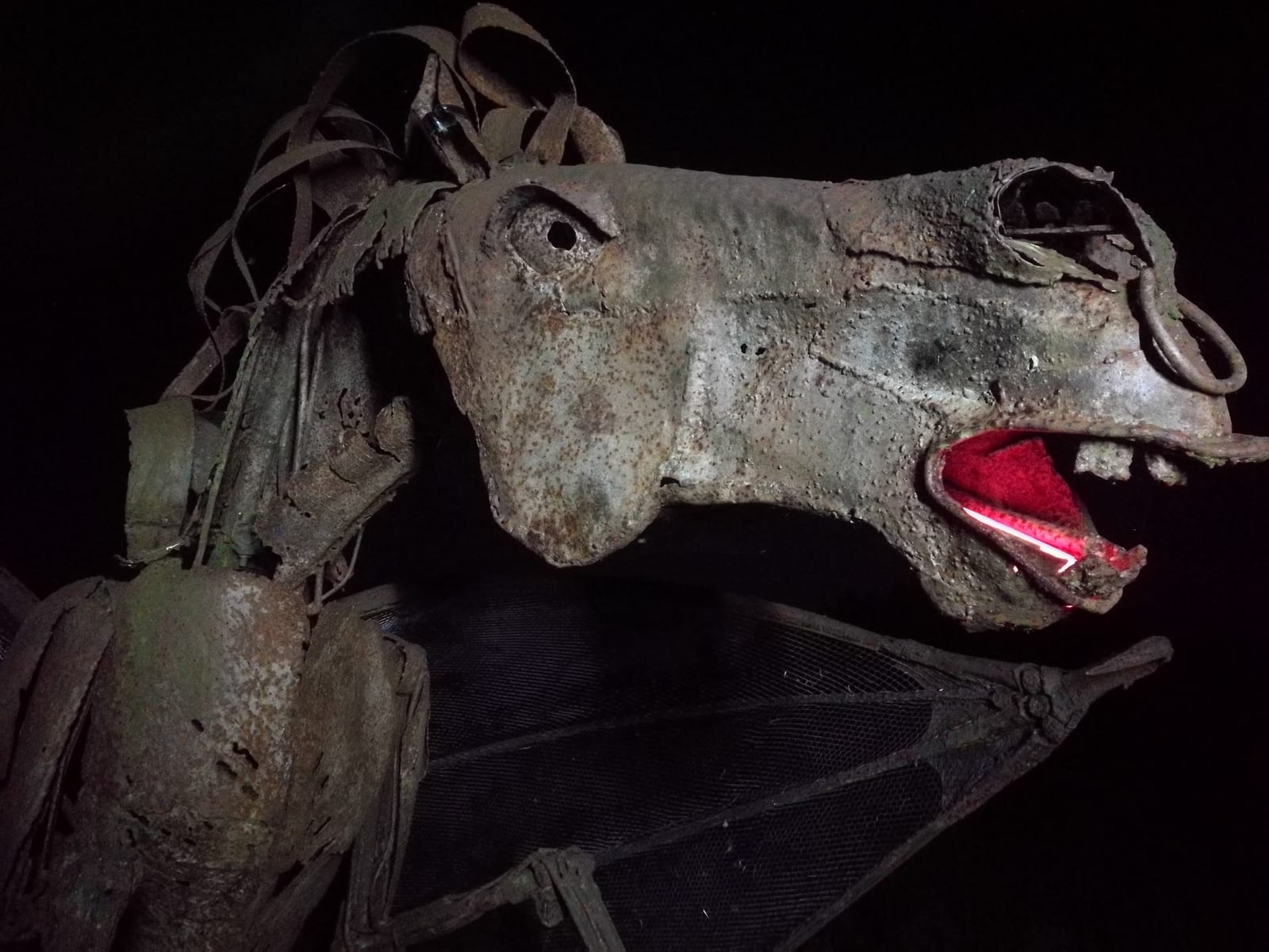 A rusty, grotesque horse-shaped metal sculpture with glowing red circles in its open mouth. The eerie figure, made of assorted metallic parts, has an unnerving expression and is captured against a dark background, enhancing its menacing appearance.