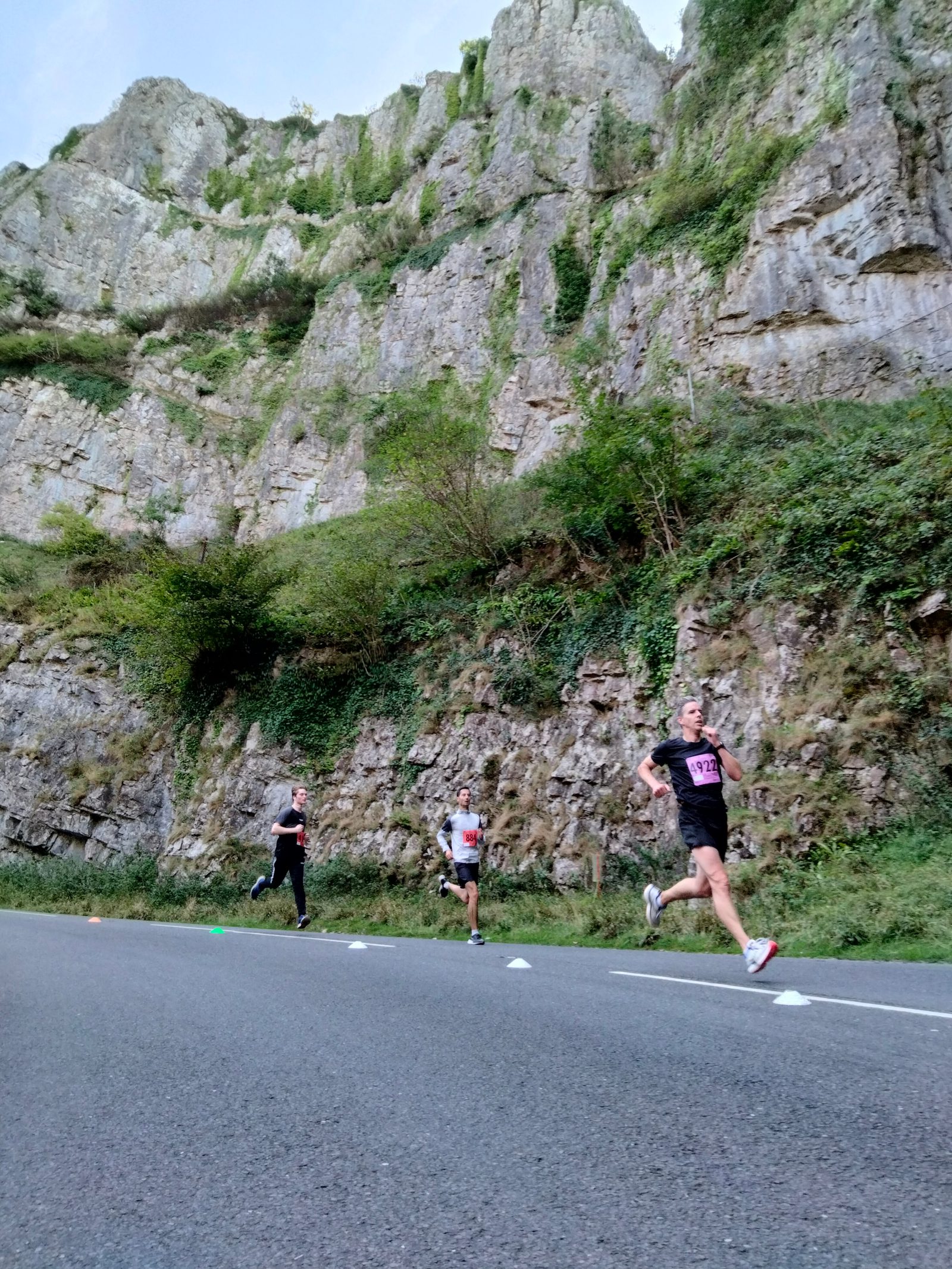 Three runners race on a paved road near a rocky, mountainous terrain with lush greenery. One runner leads considerably ahead, while the other two are closer together, trailing behind. The sky is clear, providing a picturesque backdrop to the scene.