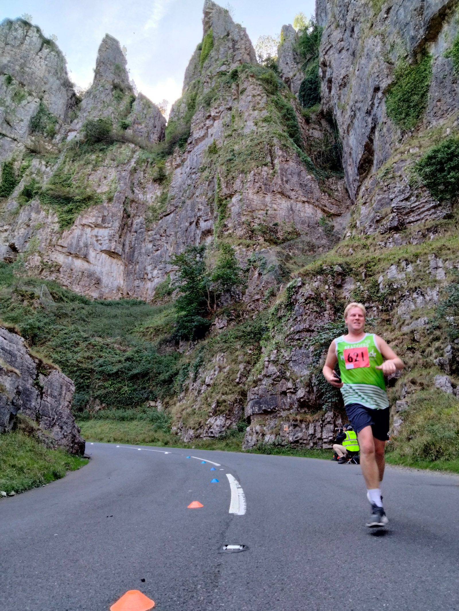 A runner wearing a green tank top and a race bib numbered 622 is running on a mountainous road. The road curves with tall, rocky cliffs covered in patches of greenery towering in the background. Orange and blue cones line the side of the path.