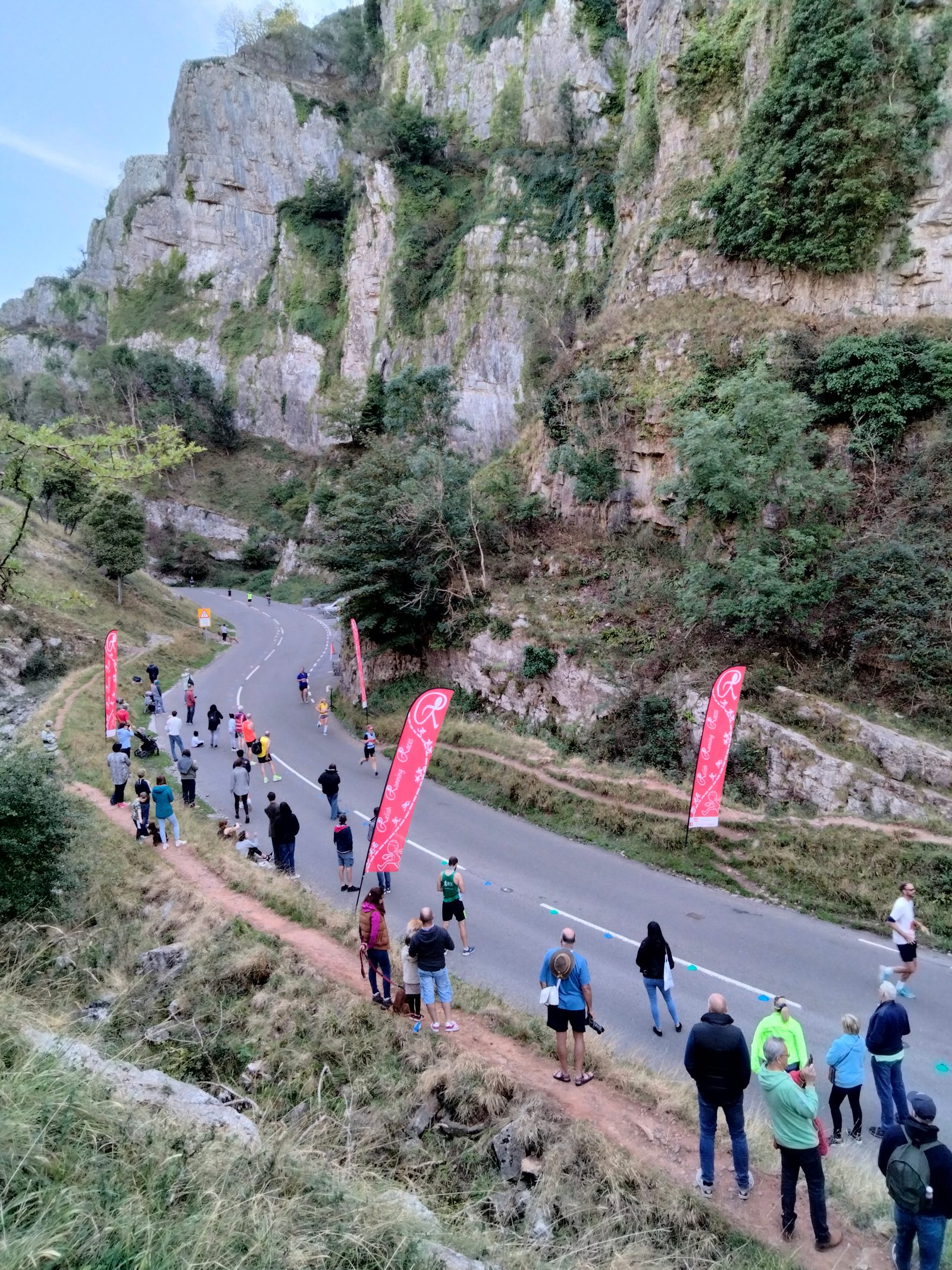 A group of runners participating in a race on a winding road through a steep, rocky gorge with lush vegetation. Spectators stand along the roadside, some holding red flags with white logos, cheering on the runners. A few cars are visible in the distance.