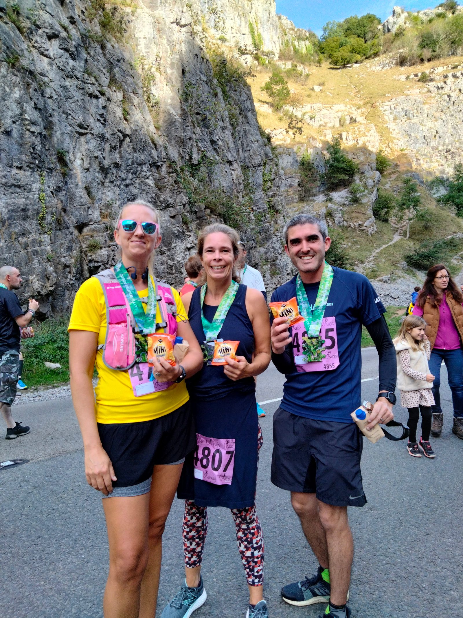 Three runners, two women and one man, wearing race bibs and medals stand smiling on a road near a rocky cliff. They hold drinks and the sun is shining. Other people and greenery are visible in the background.