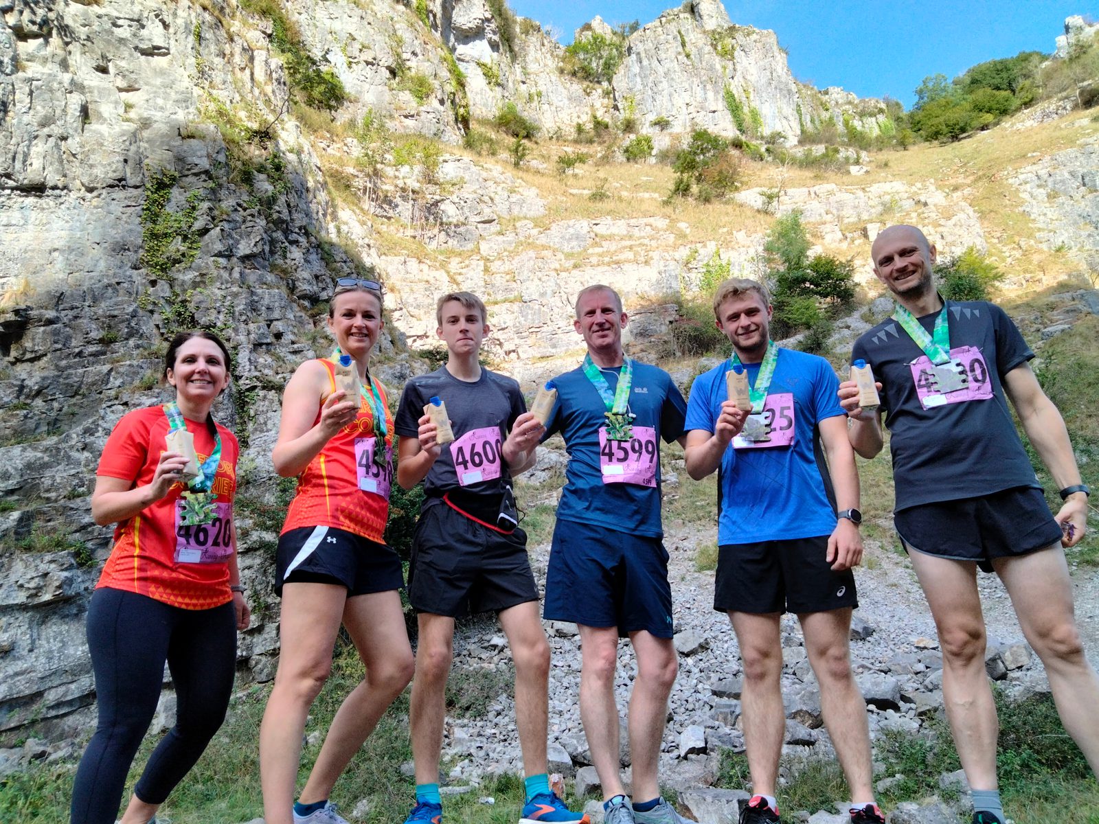 Six people in athletic attire, standing in a rocky outdoor area with medals around their necks, smiling and holding up their fingers to signify their number. They appear to have just completed a race, as indicated by the race numbers pinned to their front.