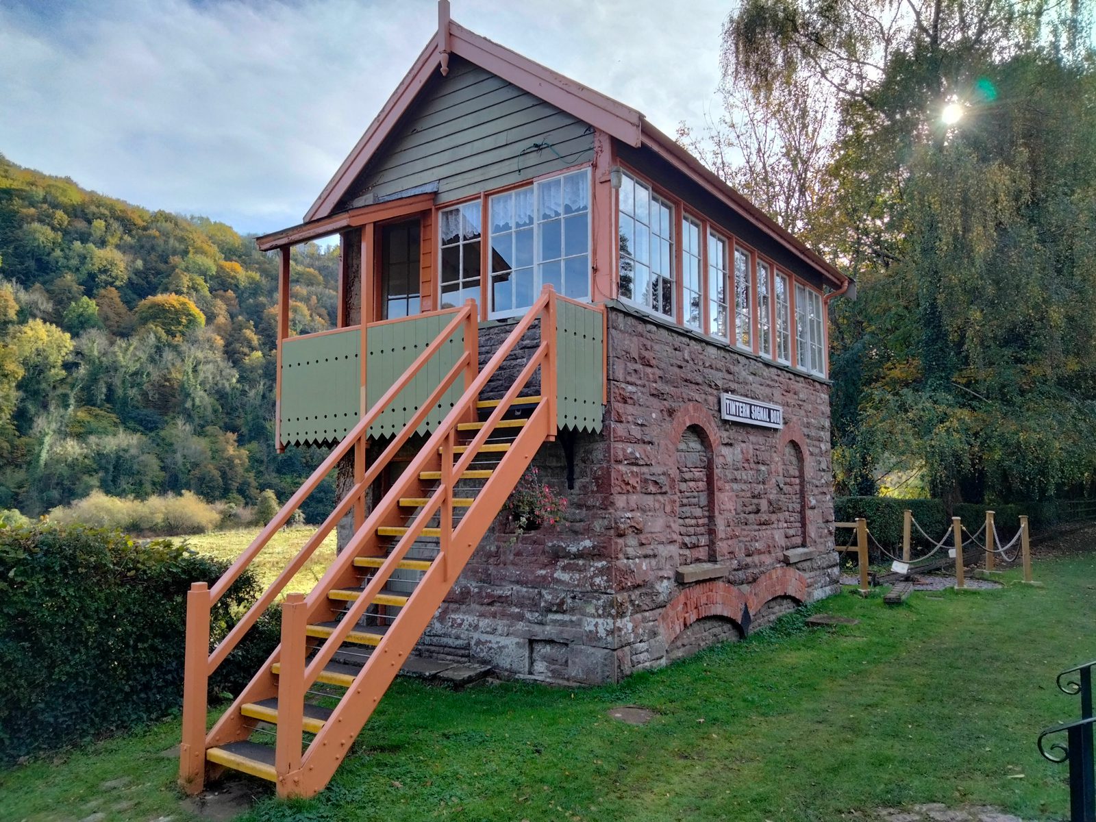 A vintage signal box stands elevated with a wooden and brick structure. A staircase leads to the entrance, with “Tintern Old Station” displayed on a sign. Surrounded by greenery, with trees in the background, the scene is tranquil and bathed in soft daylight.