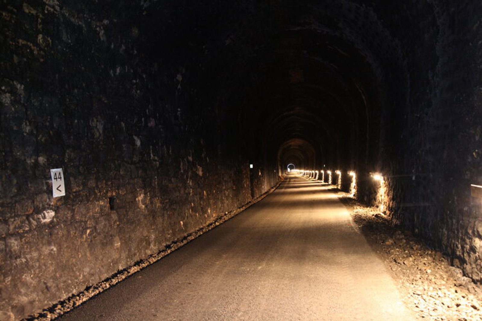 A dimly lit, paved tunnel with stone walls extending into the distance. Small lights run along one side, casting an orange glow. A small light is visible at the end of the tunnel, highlighting its length. A sign on the left shows the number 44 and an arrow.