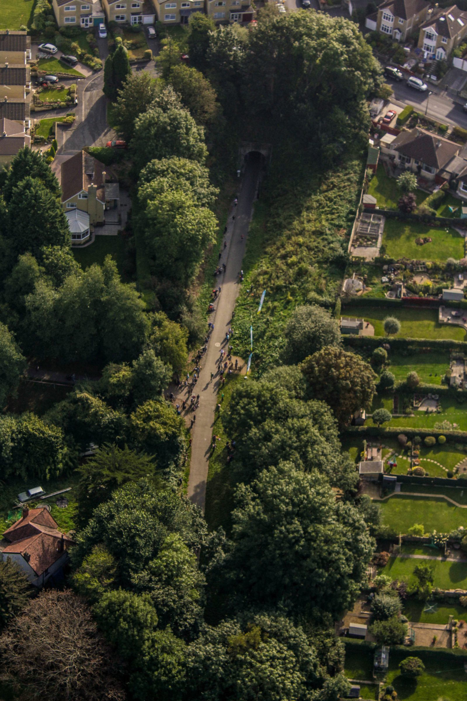 Aerial view of a road surrounded by lush greenery with people walking or cycling along it. The road leads into a tunnel at one end. Residential houses are visible on the top left, and gardens, possibly a cemetery, are on the right side of the image.