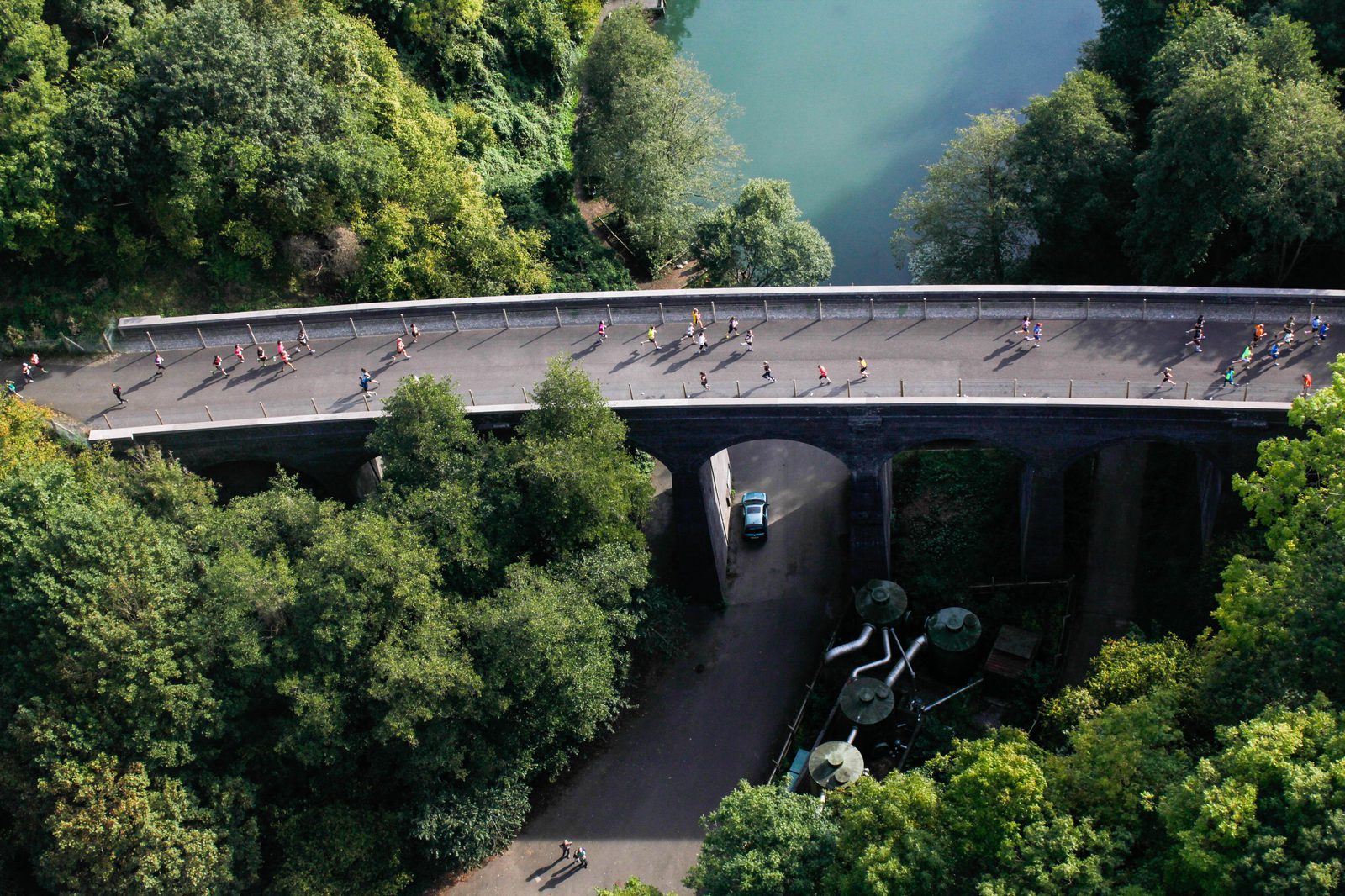 An aerial view of a group of runners on a curved bridge surrounded by lush green trees and foliage. A narrow road with a car and some structures is visible under the bridge. In the background, there's a body of water bordered by more trees.