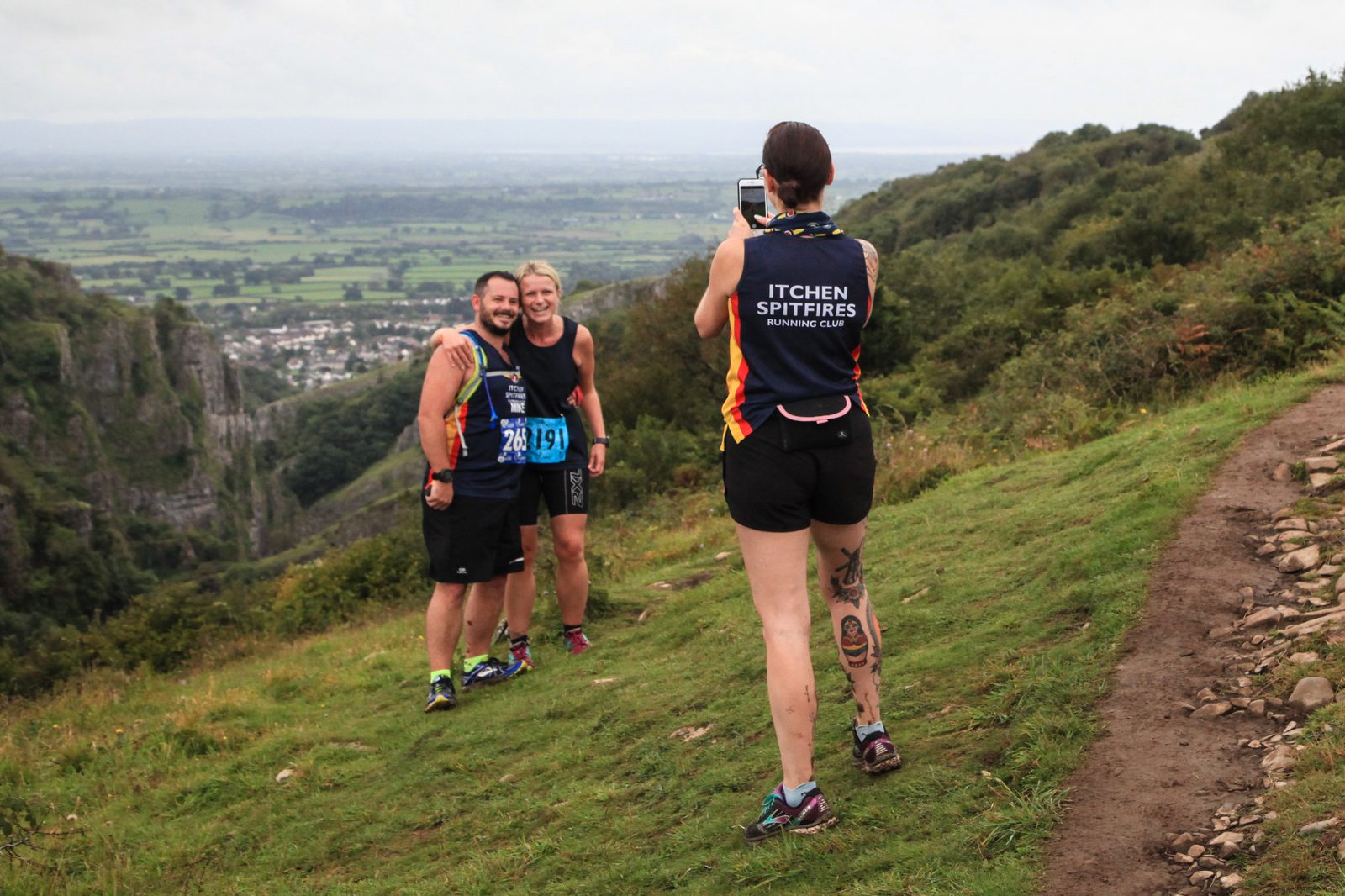 Three runners take a photo on a hill with a scenic backdrop. Two in running gear with race bibs pose, while one with "Itchen Spitfires Running Club" on a shirt takes the photo. They overlook a valley with greenery and a cloudy sky.