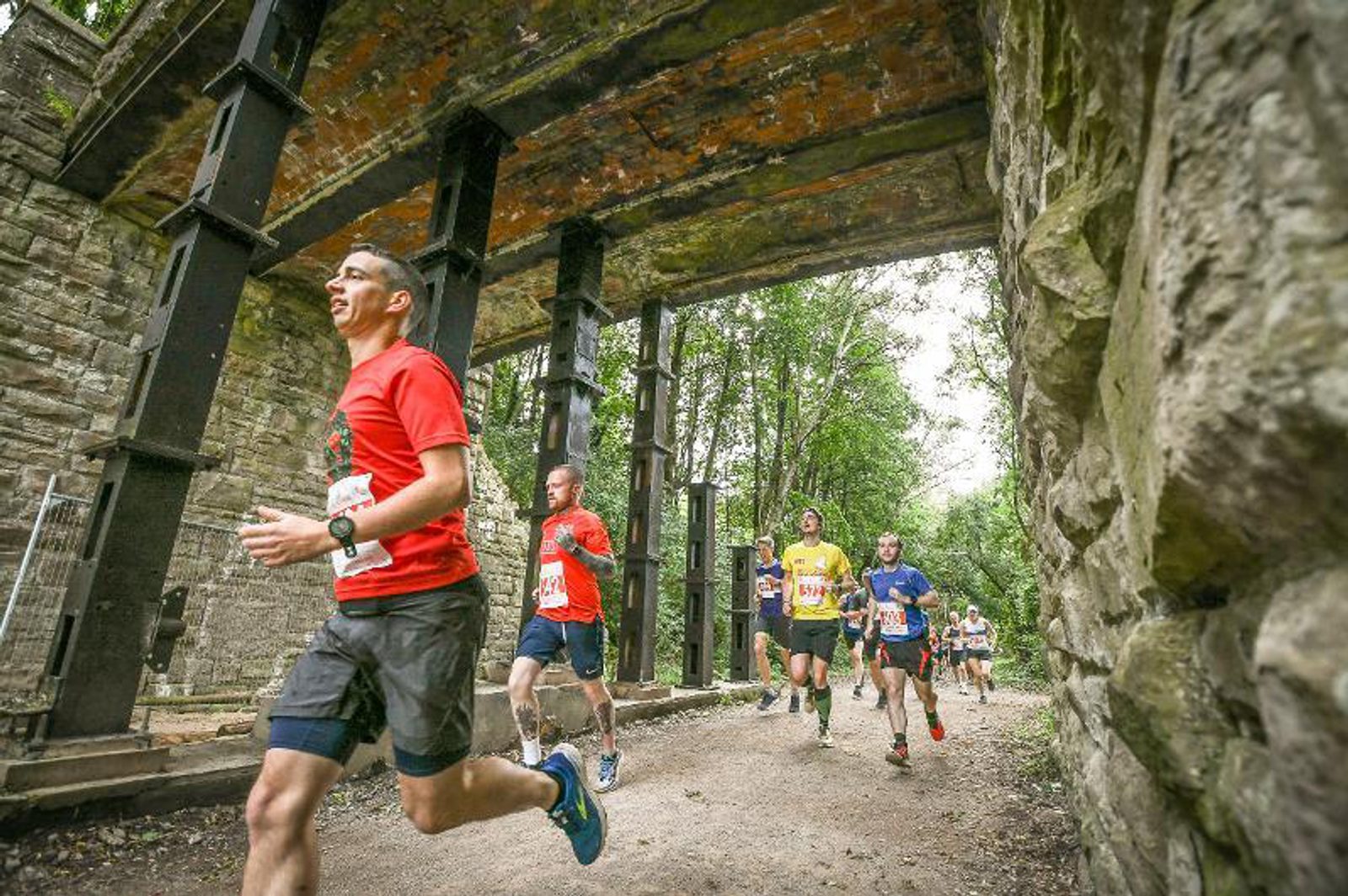 A group of runners, wearing various colored shirts and numbers, are participating in a race passing under a bridge with stone and metal supports. The background features lush green trees suggesting a forested area. The ground appears to be a dirt path.