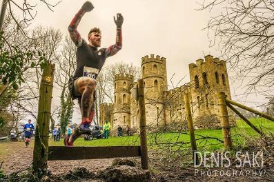 A runner in athletic gear leaps over a small hurdle during an outdoor race, with a historic castle and leafless trees in the background. Several other participants can be seen running along the path. The sky is cloudy, and the scene appears dynamic and energetic.