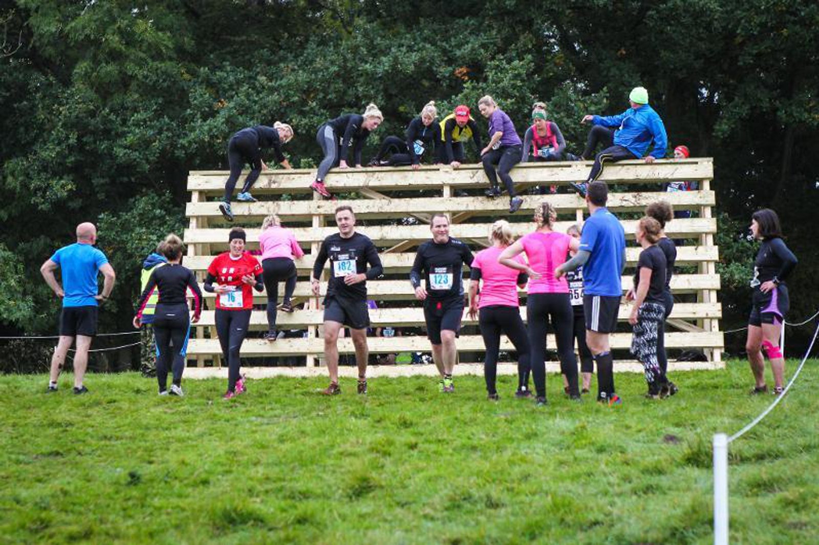Participants in athletic gear navigate a large wooden climbing obstacle during an outdoor race event, with some individuals helping each other over the structure. Others are waiting or moving at the base, and the background is filled with trees.