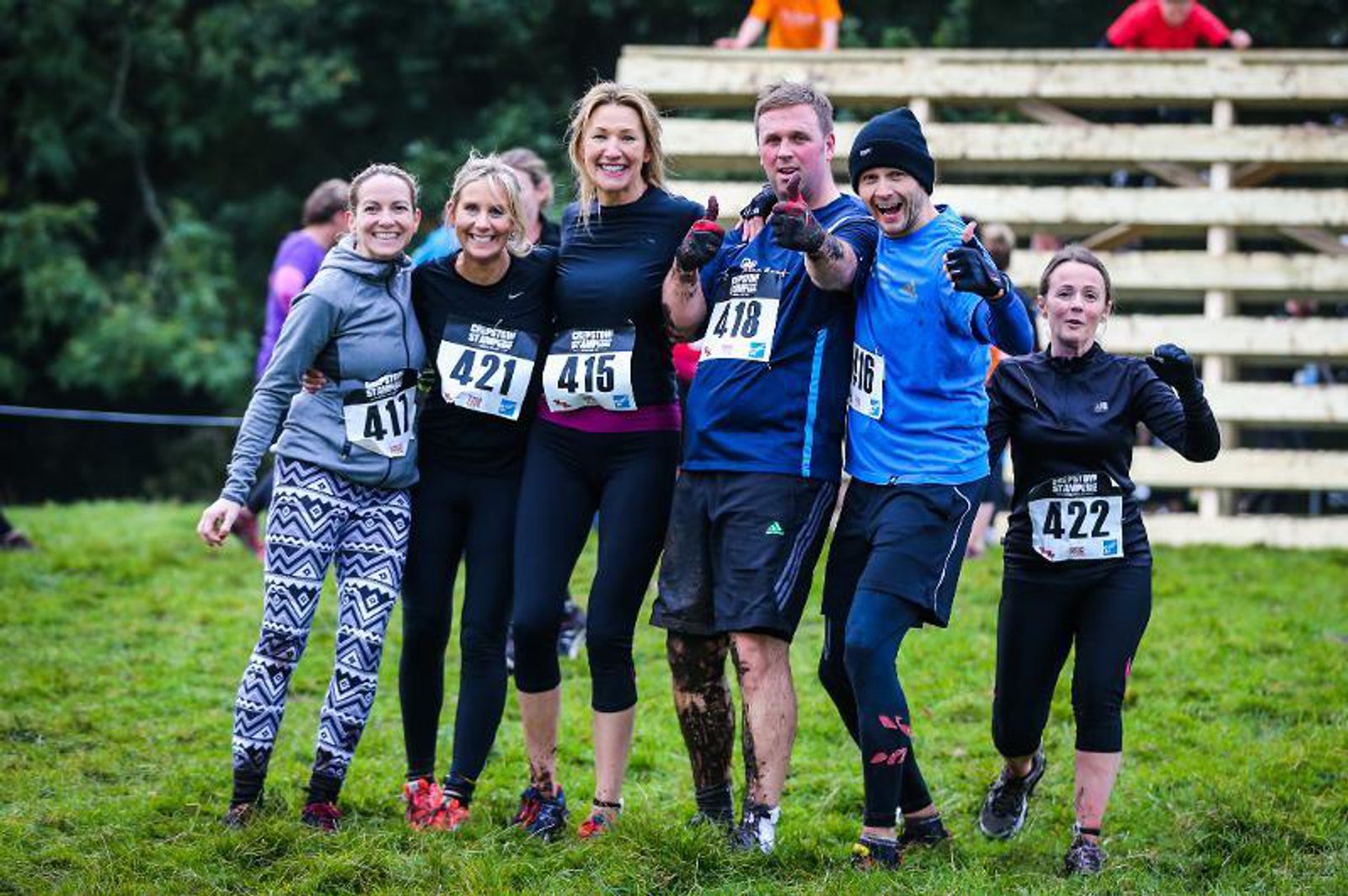 A group of six enthusiastic runners, wearing race bibs numbered 417, 421, 415, 418, 416, and 422, pose together and smile for the camera. They are outdoors in a grassy area with an obstacle course structure in the background. The runners appear muddy and happy.