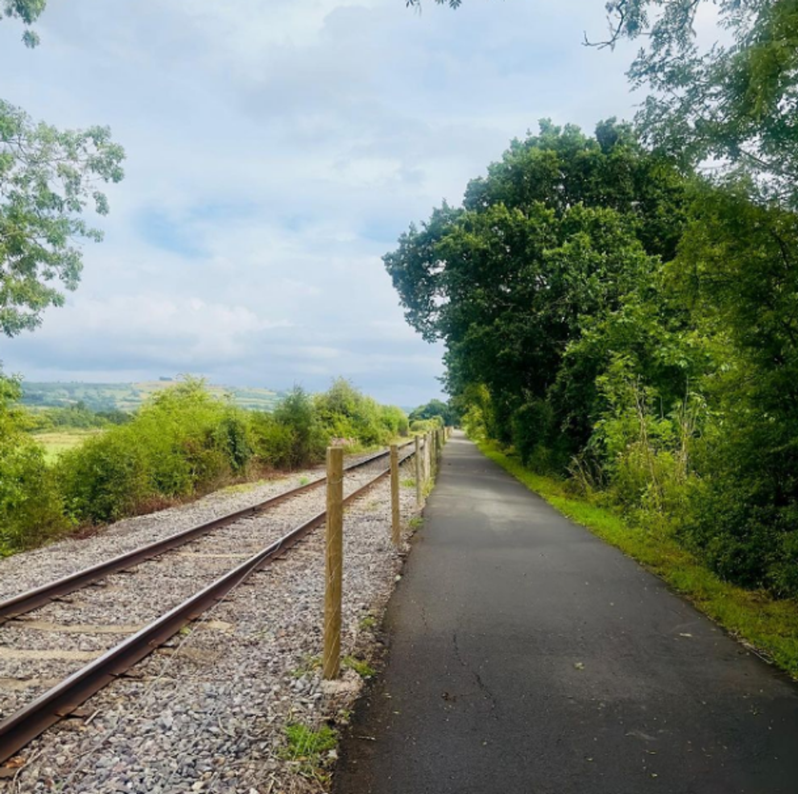 A tranquil scene featuring a paved path running parallel to a railway track surrounded by lush greenery. The sky is partially cloudy, adding to the serene atmosphere. The area is lined with trees and bushes, creating a peaceful, natural setting for walking or biking.