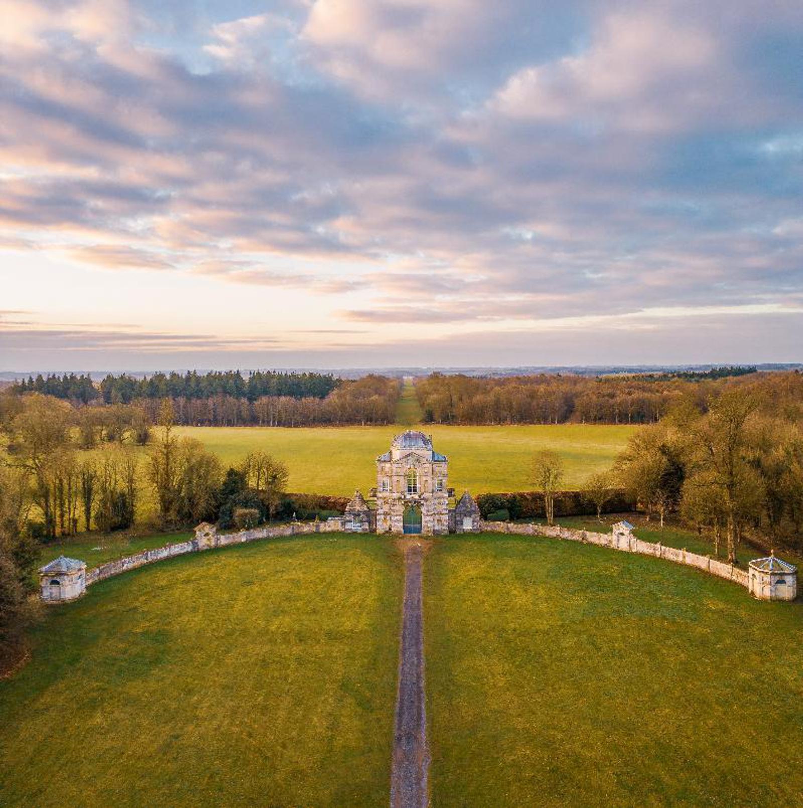 Aerial view of an ornate stone gatehouse and path leading to it, surrounded by expansive green fields and bordered by trees. The sky is partly cloudy, with the sun casting warm light over the landscape.