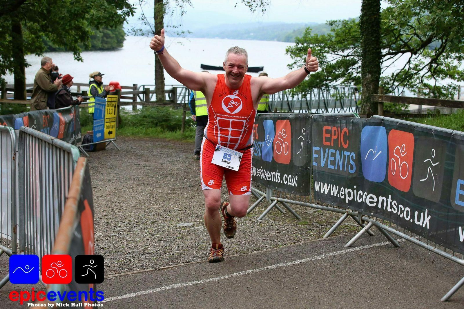 A runner dressed in a red sports outfit and bib number 65 crosses the finish line with arms raised in victory. Event banners for Epic Events line the pathway, and a lake can be seen in the background. Spectators are visible near the finish area.
