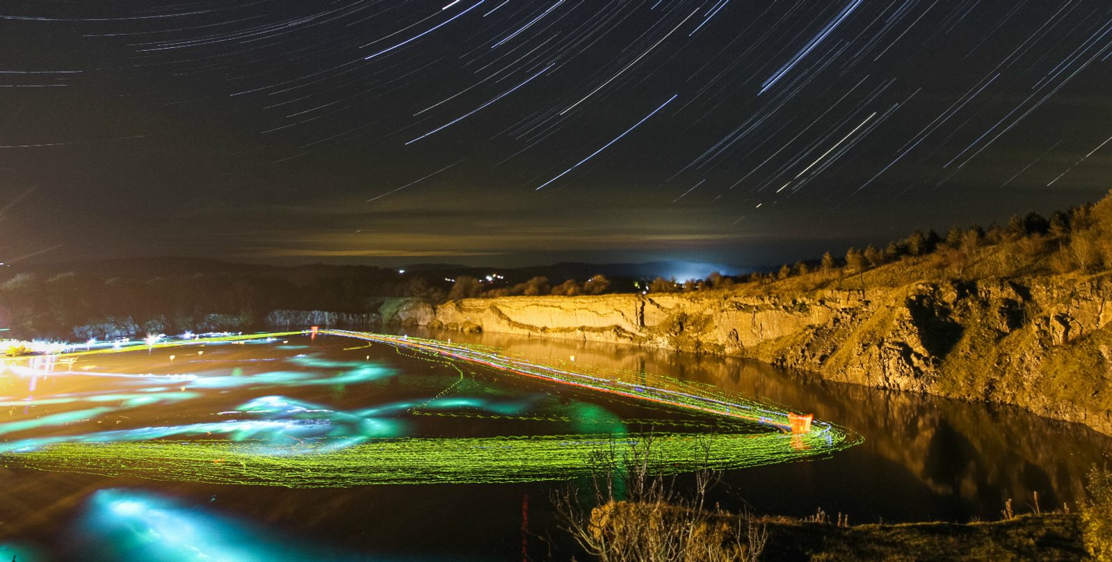 A night sky with star trails over a calm lake surrounded by rocky cliffs and trees. Colorful light trails create an intricate pattern on the water's surface, reflecting the ambient glow. The scenery is illuminated by a distant light source, enhancing the tranquil atmosphere.