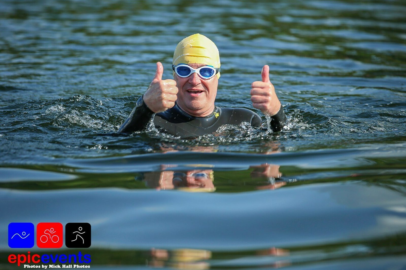 A swimmer wearing a yellow swim cap, blue goggles, and a black wetsuit is in the water giving two thumbs up. The person appears to be smiling. The water is calm, and there are graphics and text at the bottom left corner that say "epic events.