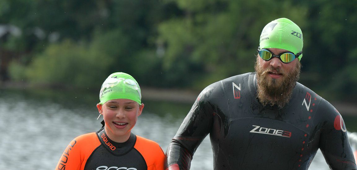 Two swimmers, wearing bright green swim caps and black wetsuits with orange accents, emerge from a body of water. Both are smiling, with the individual on the right sporting a beard and reflective sunglasses. Trees and water are visible in the background.