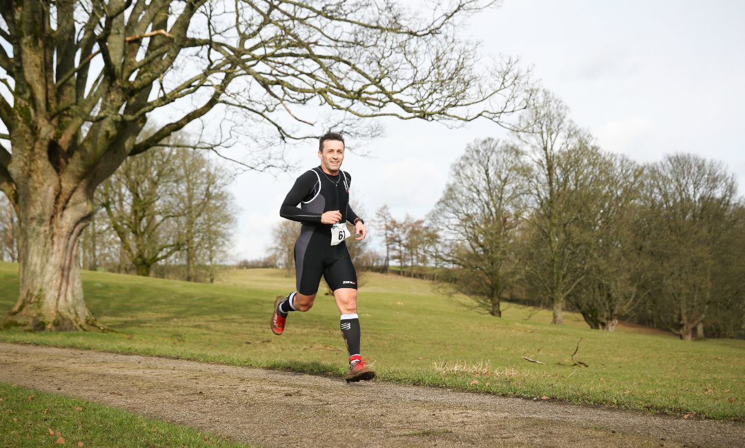 A runner wearing black athletic gear and red running shoes jogs on a path in a park. Bare trees and a grassy field fill the background under a cloudy sky. The runner wears a race bib with the number "14.