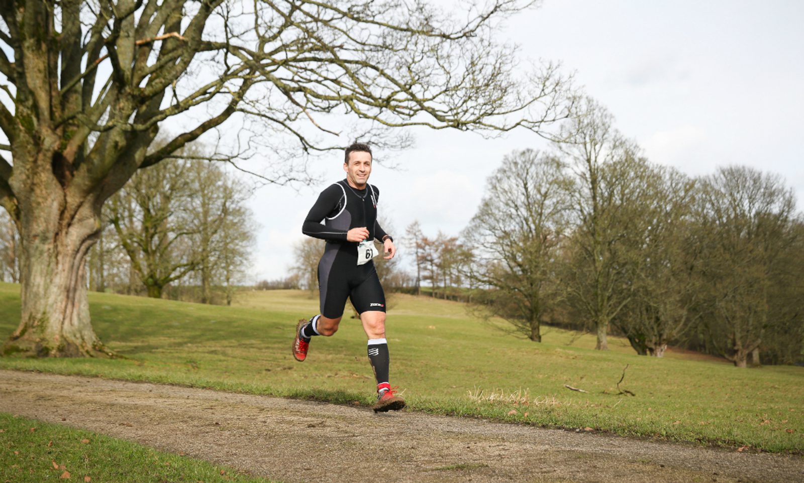 A runner wearing black athletic gear and red running shoes jogs on a path in a park. Bare trees and a grassy field fill the background under a cloudy sky. The runner wears a race bib with the number "14.