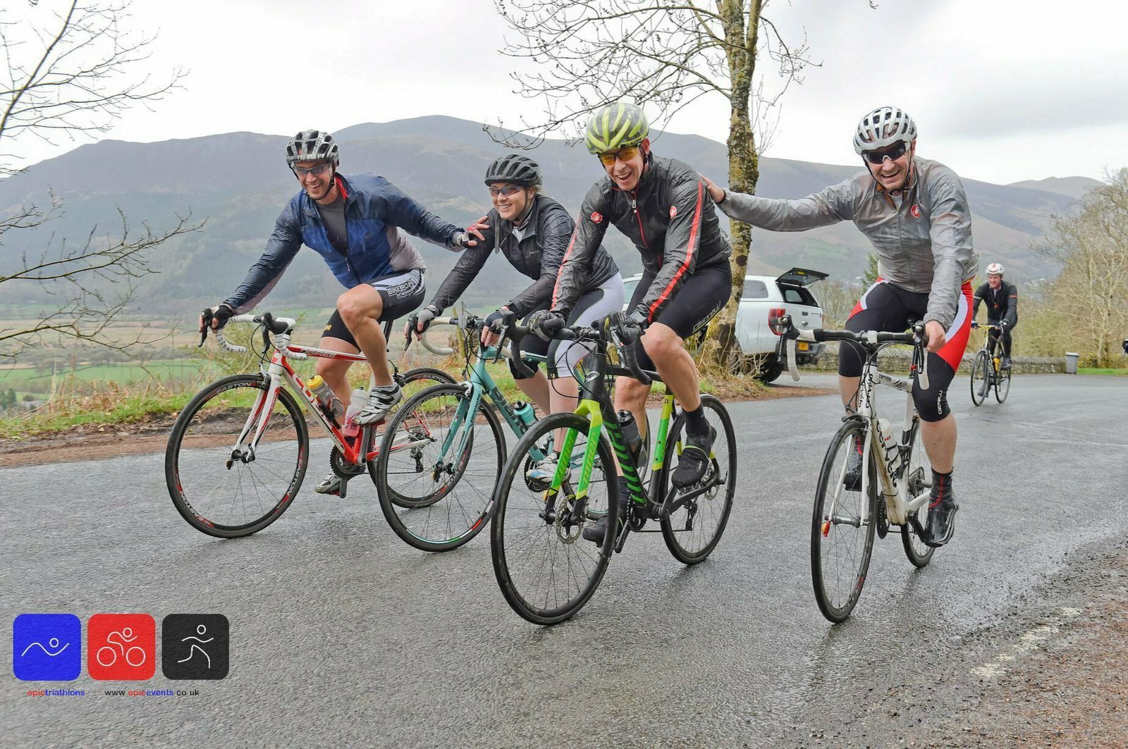 Four cyclists in rain gear ride on a wet, paved road in a hilly, wooded area. They are smiling and extending arms to each other, enjoying the camaraderie. Two icons at the bottom left depict a figure swimming, a cyclist, and a runner. A car is parked in the background.