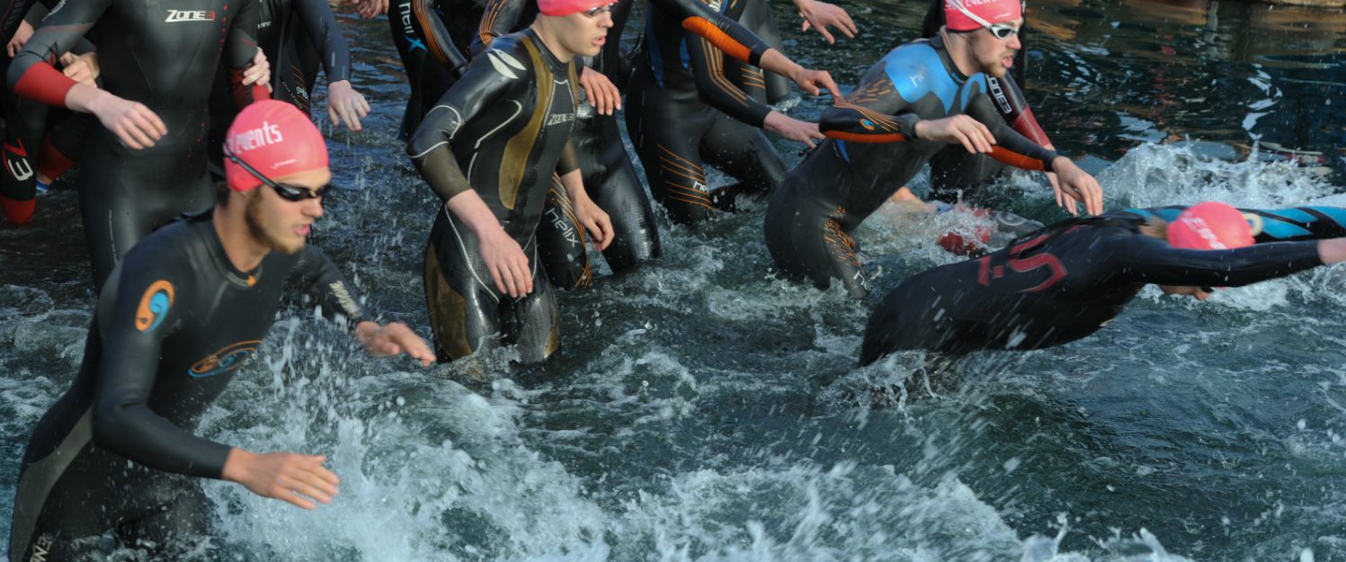 A group of triathletes in wetsuits and pink swimming caps is seen starting the swim portion of a triathlon. They are splashing into the water with determined expressions, creating waves and foam as they begin the race.