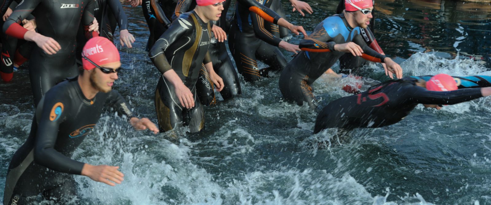 A group of triathletes in wetsuits and pink swimming caps is seen starting the swim portion of a triathlon. They are splashing into the water with determined expressions, creating waves and foam as they begin the race.