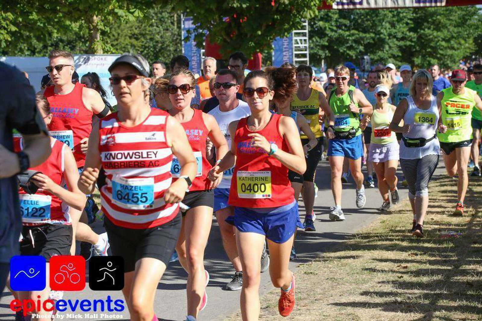 Runners participating in a race, with some wearing bib numbers and others in bright athletic gear. The event is hosted by "Epic Events," as indicated by a logo at the bottom left corner. The runners are focused and moving along a tree-lined path on a sunny day.