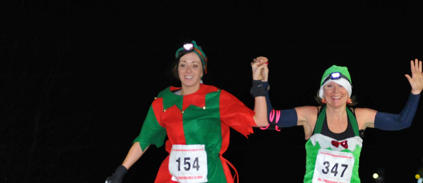 Two women are running in a nighttime race, smiling and holding hands. They are wearing festive costumes, including headlamps. The woman on the left is dressed as an elf and the woman on the right is wearing a Christmas-themed outfit. Both have race numbers pinned on.