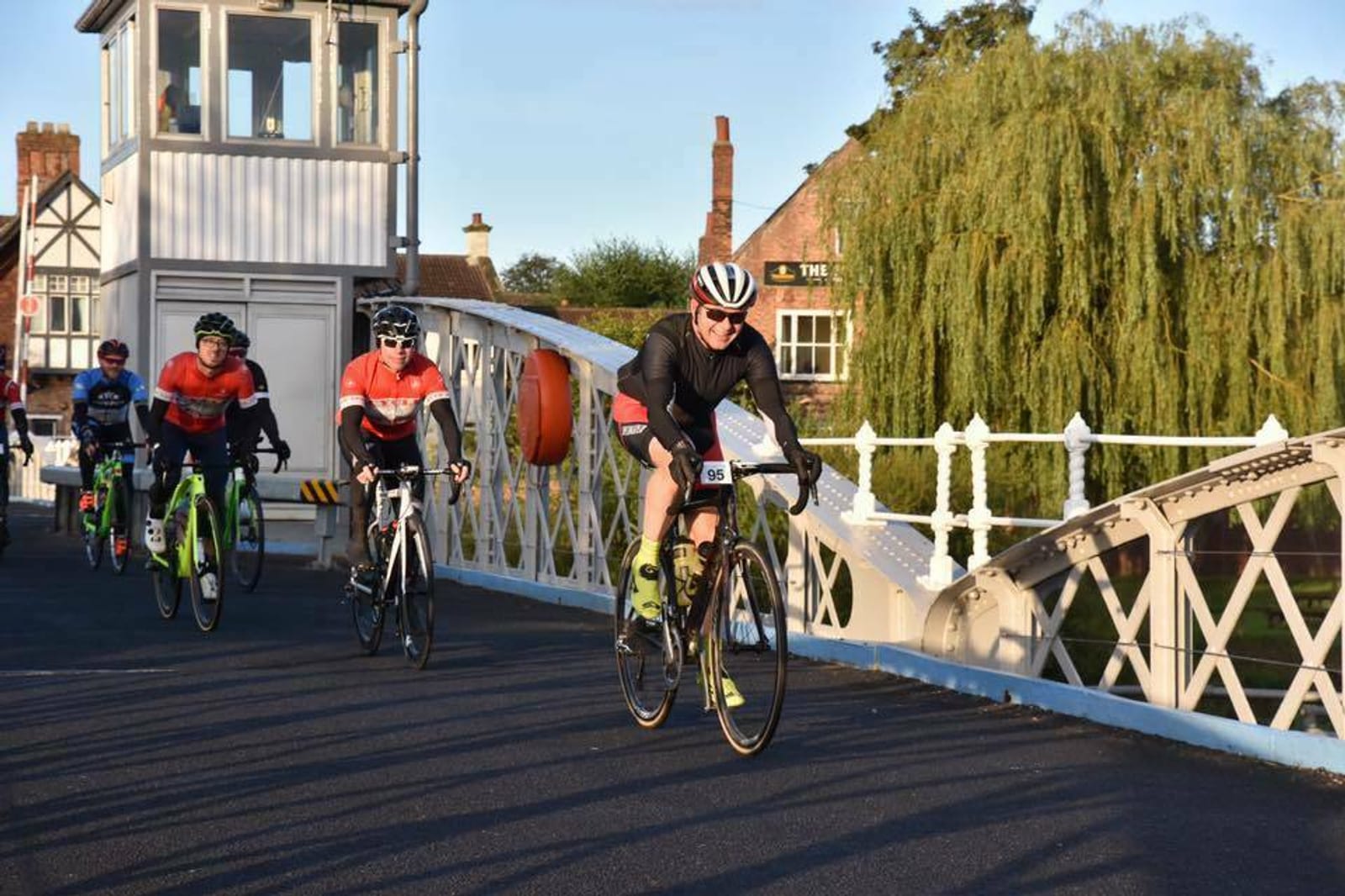 A group of cyclists is riding across a bridge. In the foreground, a cyclist with a black jersey and helmet is in the lead. Other cyclists wearing red and black jerseys follow behind. The bridge has white railings, and a green tree is visible in the background.