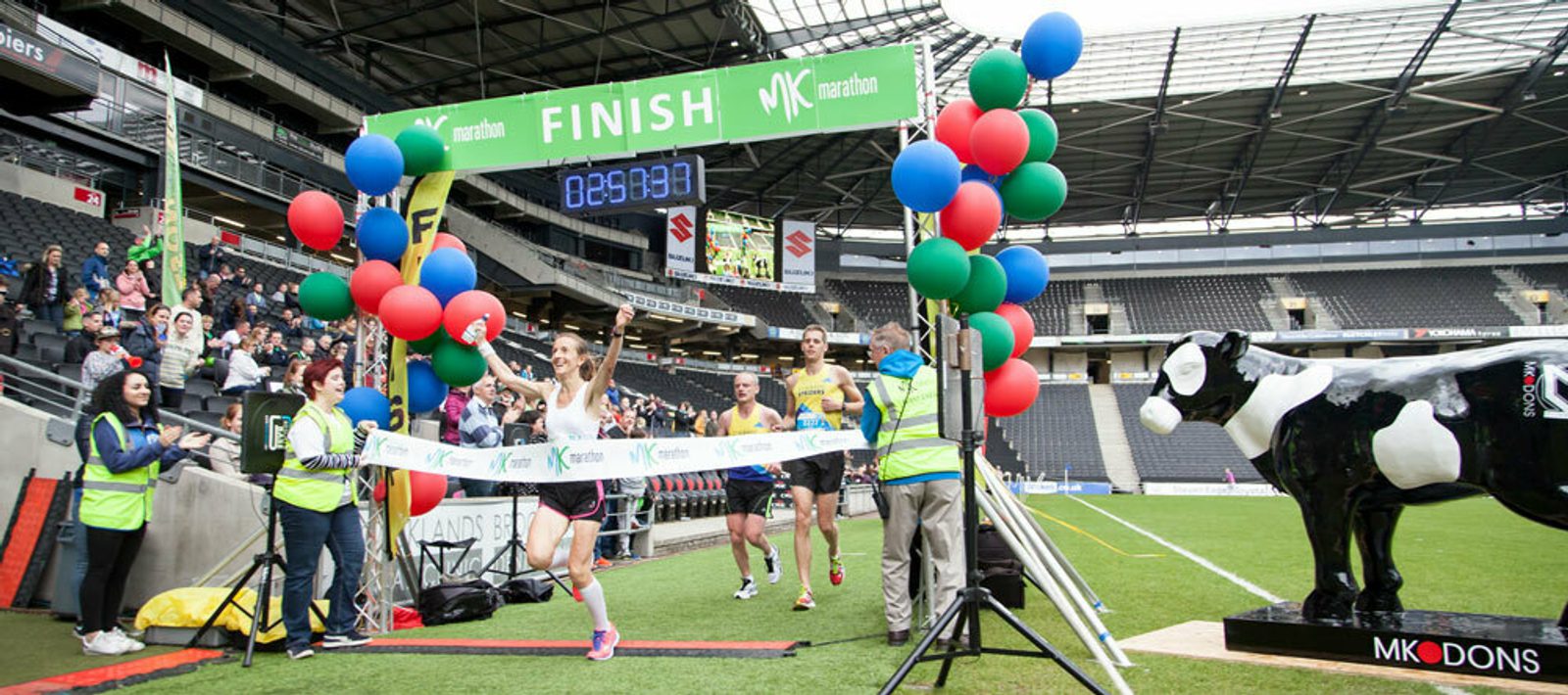 A runner crosses the finish line at a marathon in a stadium, raising their arms in victory. Other runners follow behind. The finish line is adorned with colorful balloons and a large cow statue stands to the right. Spectators and event staff cheer in the background.
