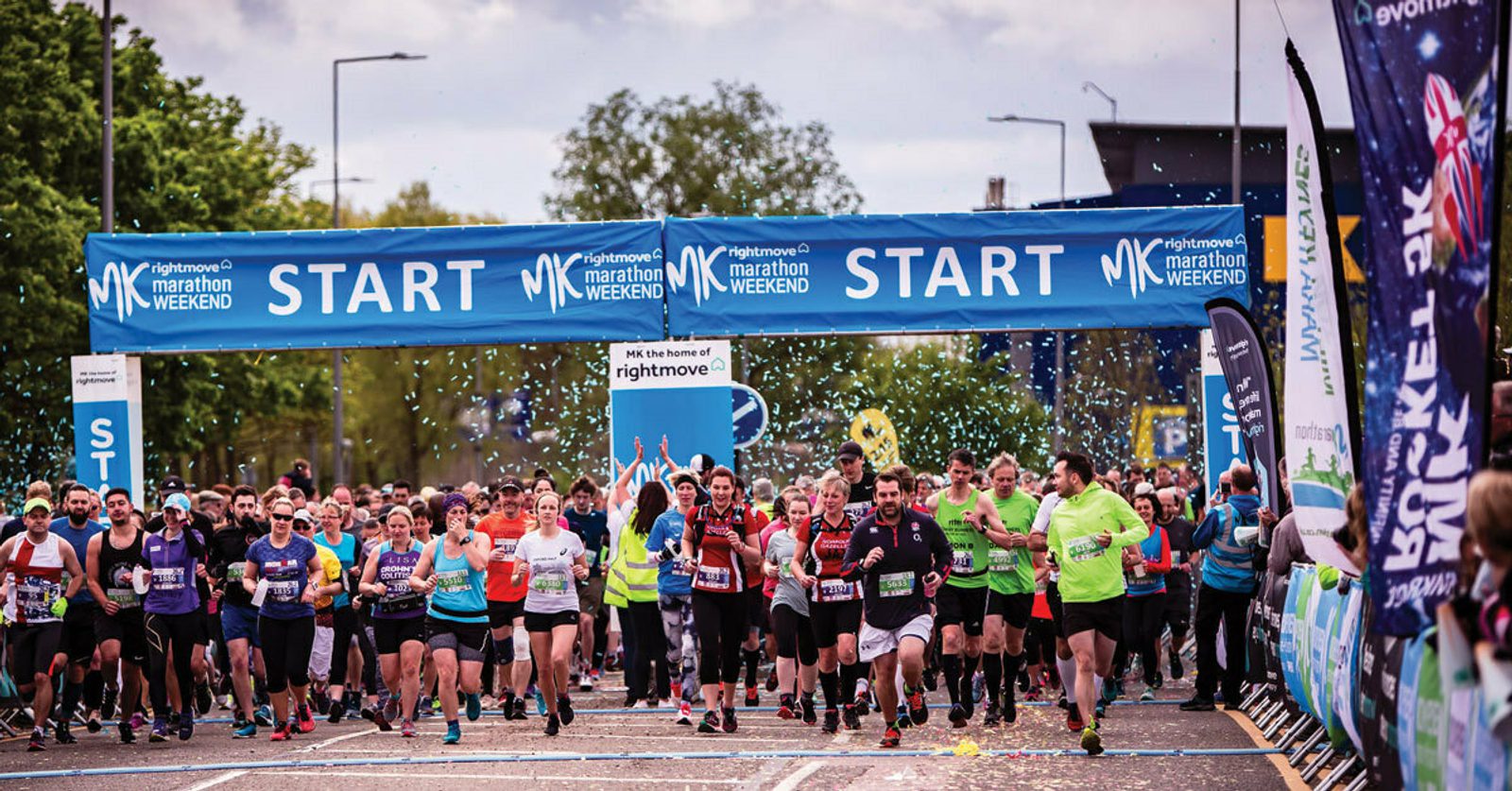 A large group of runners crosses the starting line at the MK Marathon Weekend event. They are surrounded by spectators and blue banners that read "START" and "MK Marathon Weekend." Confetti fills the air, and the runners are dressed in a variety of athletic gear.