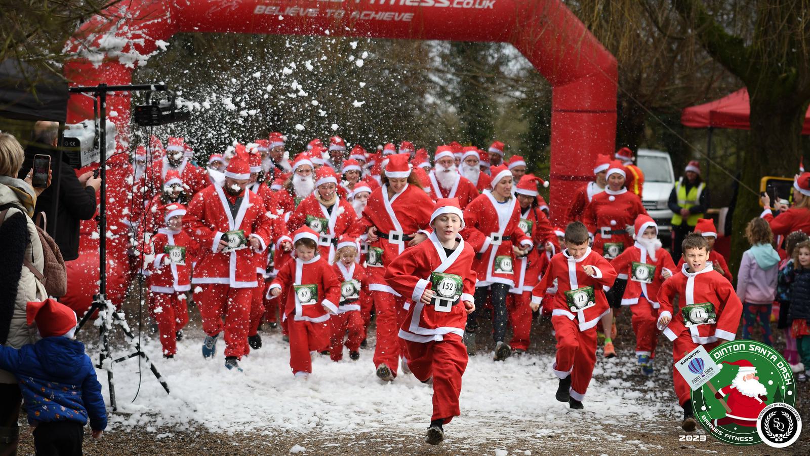 A large group of people dressed in Santa Claus costumes running through a start line arch at an outdoor event. Spectators and festive decorations are visible in the background, with artificial snow creating a wintry atmosphere.