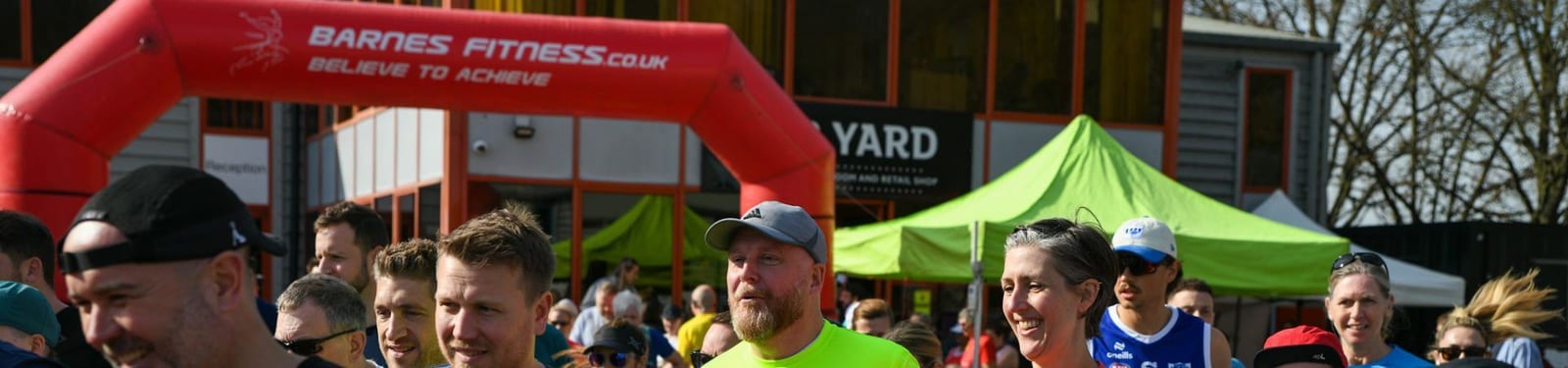 A group of runners gather at the start line under a red inflatable arch that reads "Barnes Fitness. Believe to Achieve," with green tents and a building visible in the background.