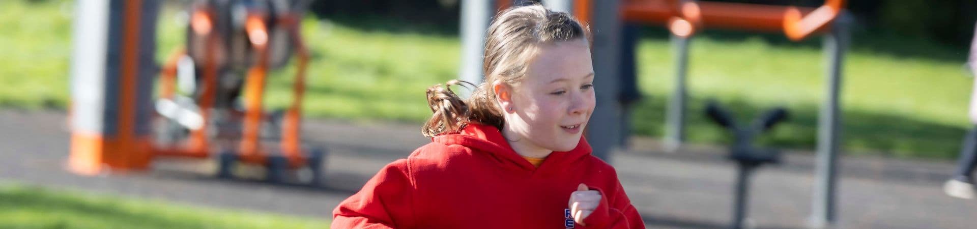 A young girl in a red hoodie runs outdoors in a park or playground, with exercise equipment and green grass visible in the background.