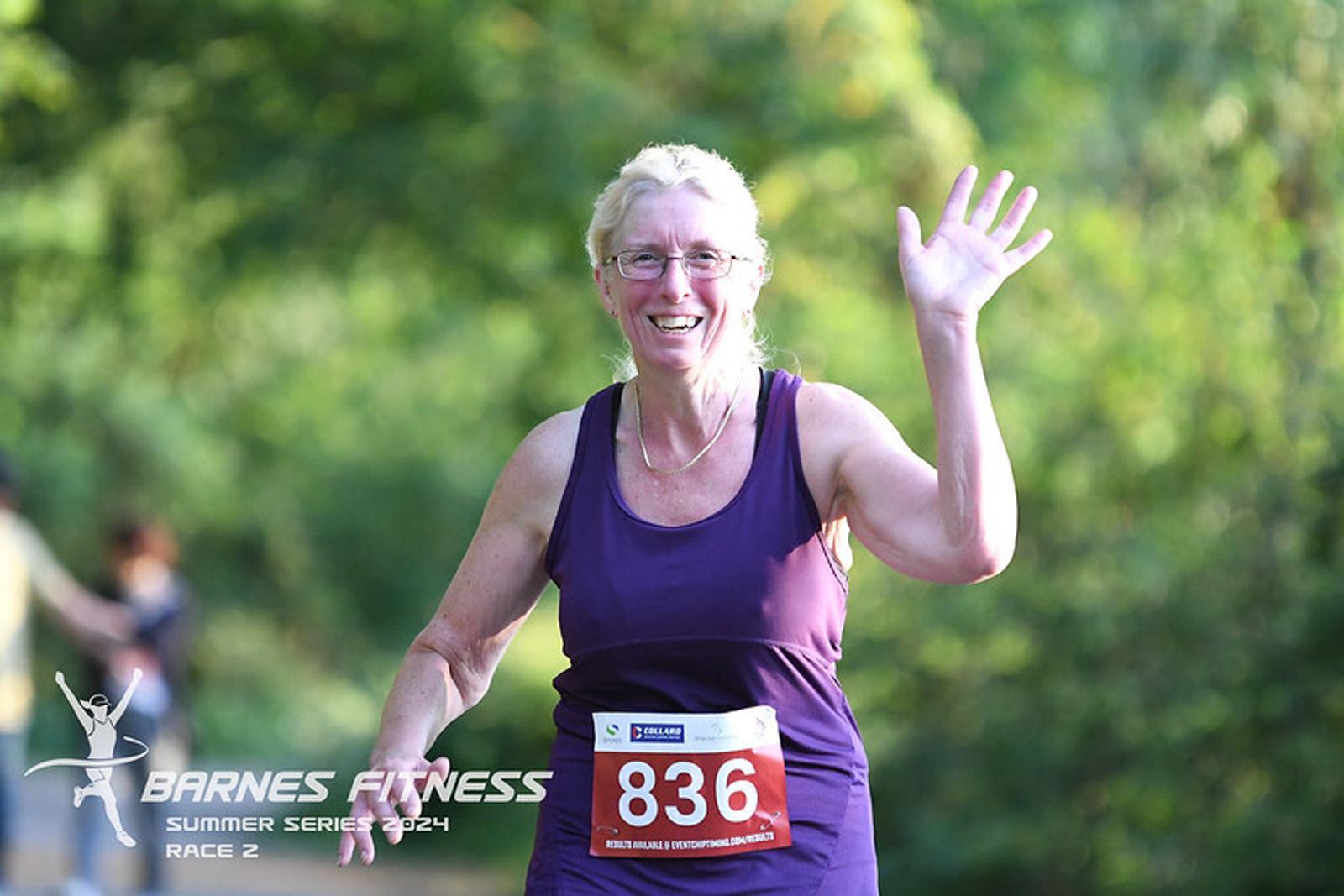 An older adult with a race number 836, wearing a purple tank top, smiles and waves while running in a park setting. The image has a "Barnes Fitness Summer Series 2024 Race 2" logo.