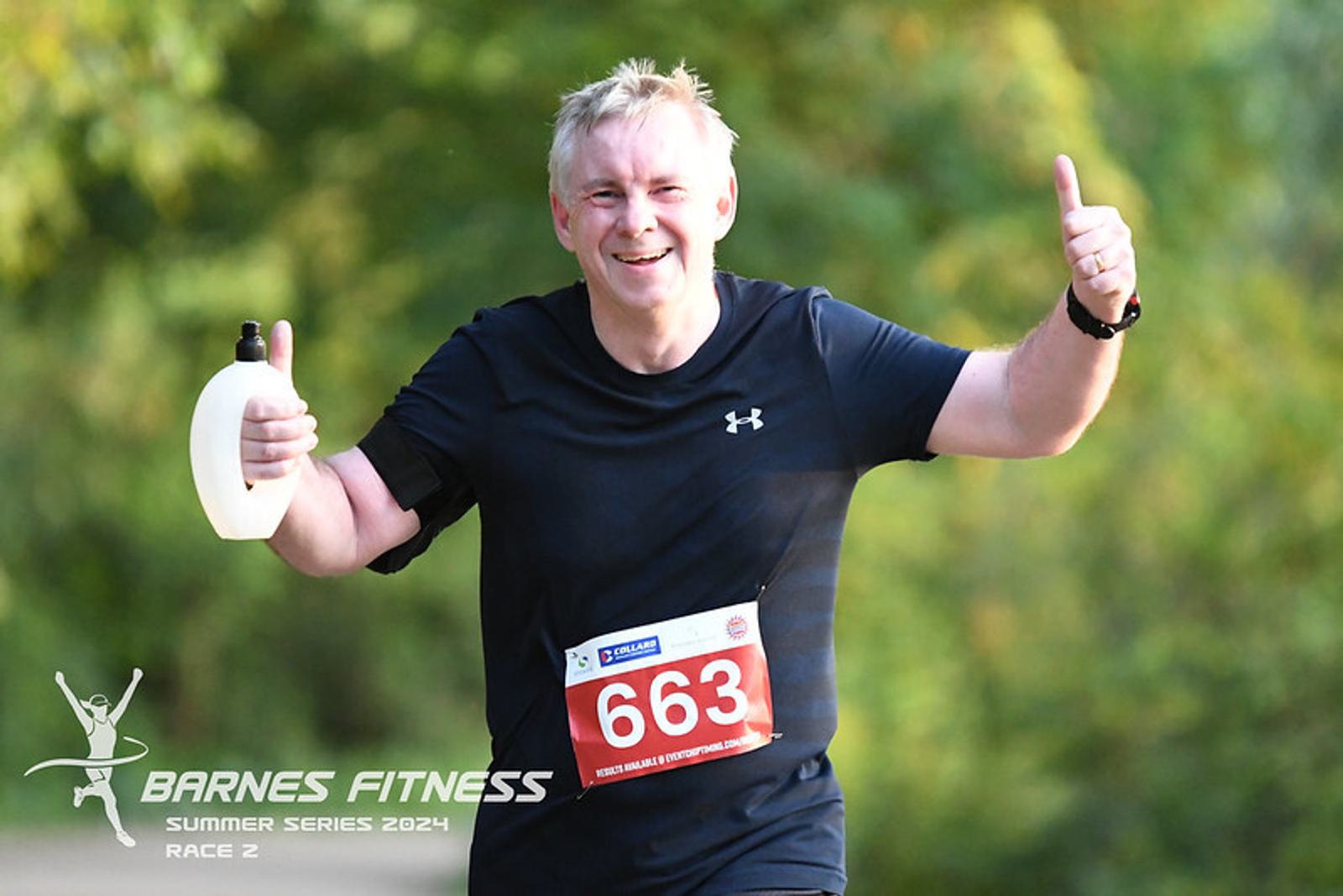 A man in a black running outfit is participating in a race, smiling and giving a thumbs-up with both hands. He's holding a small water bottle in his left hand. His race number is 663. Trees and foliage are visible in the background.