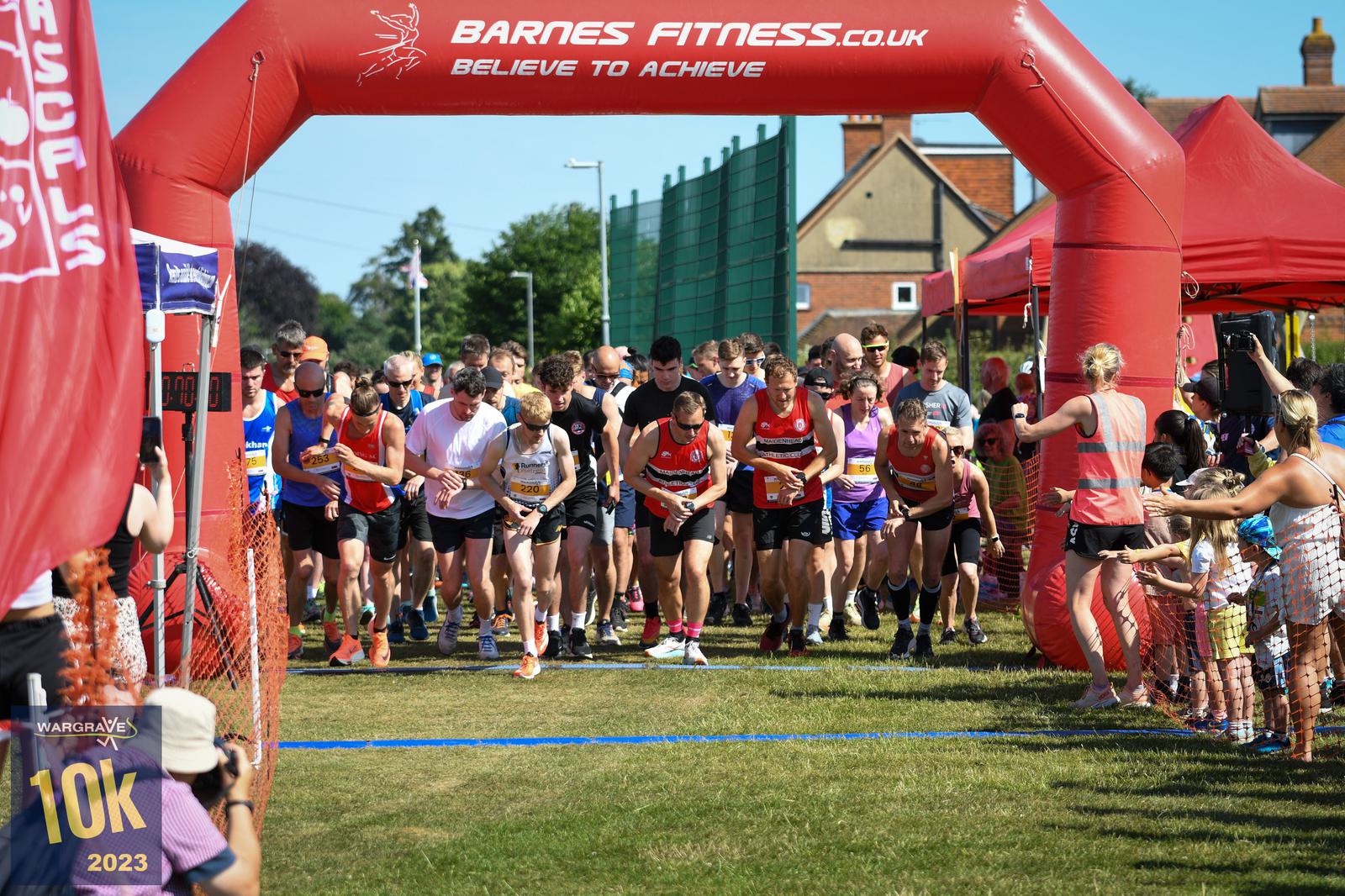 A diverse group of runners begins a race under a red inflatable arch that reads "Barnes Fitness" and "Believe to Achieve." Spectators line the sides of the grassy path, cheering them on. A sign with "Wargrave 10K 2023" is visible.