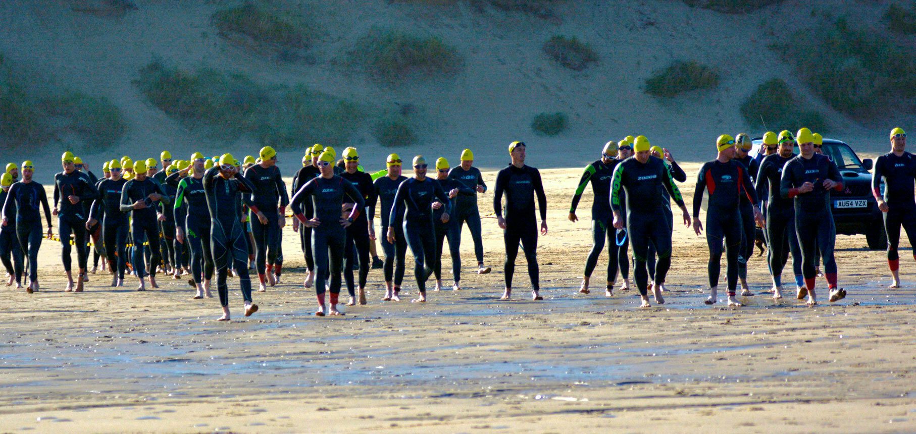 A large group of people wearing wetsuits and yellow swim caps walk across a sandy beach toward the water. The backdrop of the image includes sand dunes and a parked car. The sky appears clear.