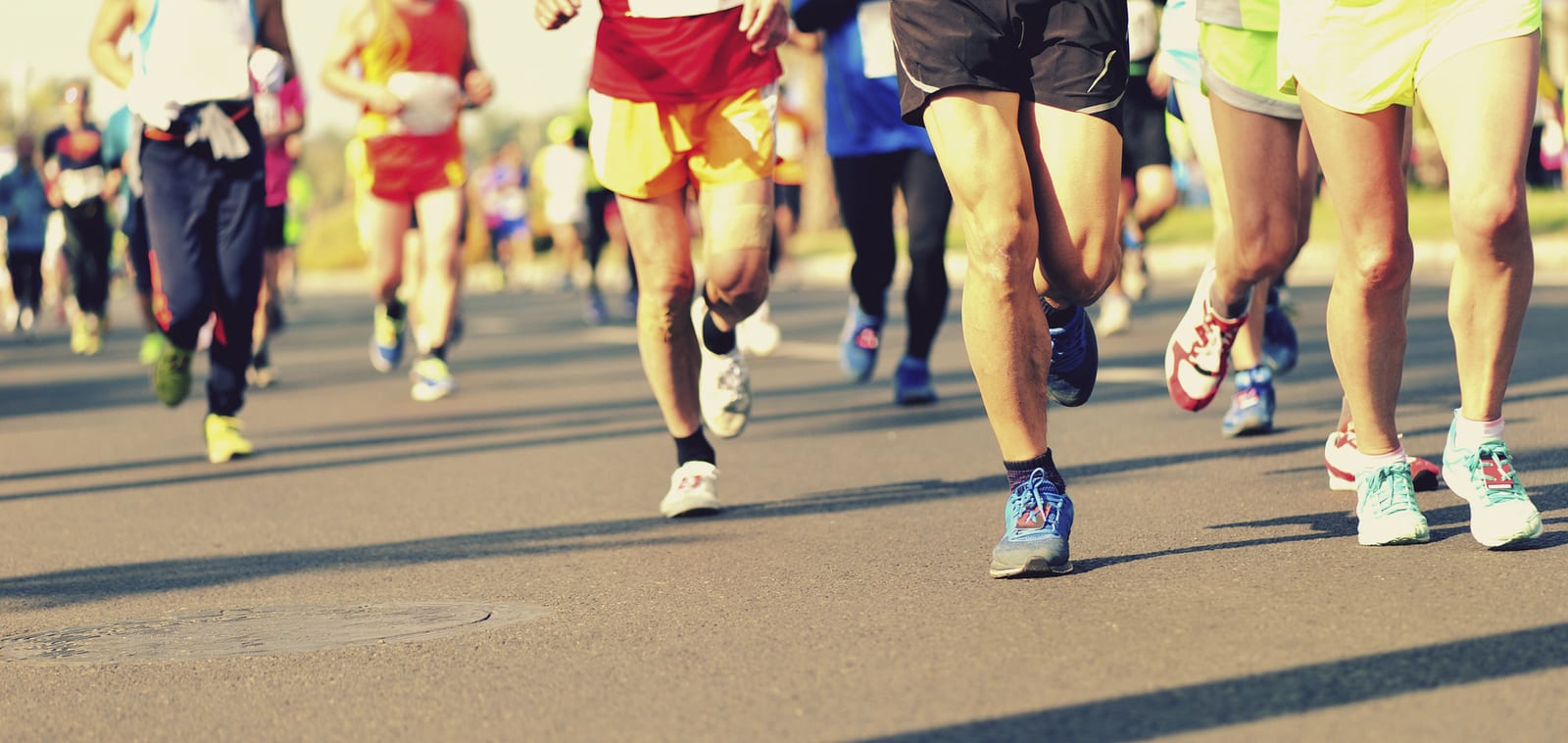 A group of runners wearing various colorful athletic outfits are running on a paved road during a race. The image captures their legs and lower bodies with a clear view of their different running shoes. The background is slightly blurred.