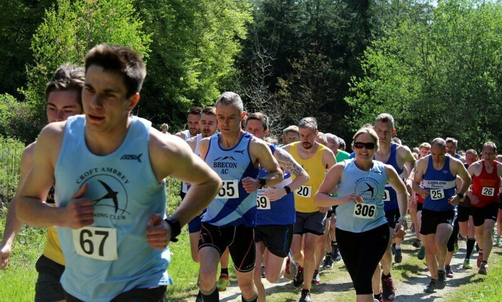 A group of runners participating in an outdoor race on a sunny day. The runners, wearing various athletic gear and race bibs, are focused and determined, running through a forested path surrounded by greenery. The foreground runner wears bib number 67.