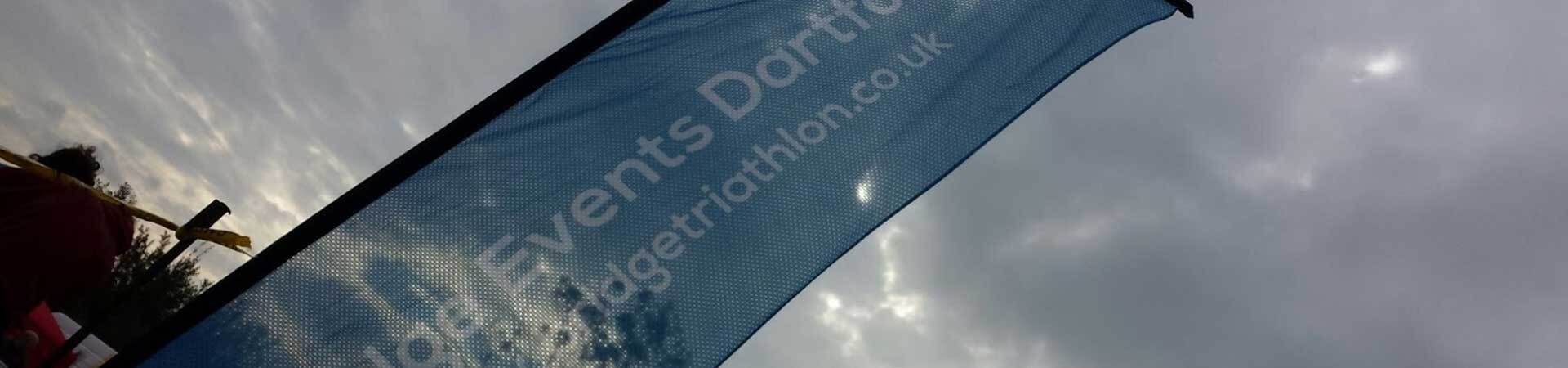 A blue event banner with white text stands against a cloudy sky. The visible text includes “Events Dart… getriathlon.co.uk.” Yellow tape and trees appear in the background.