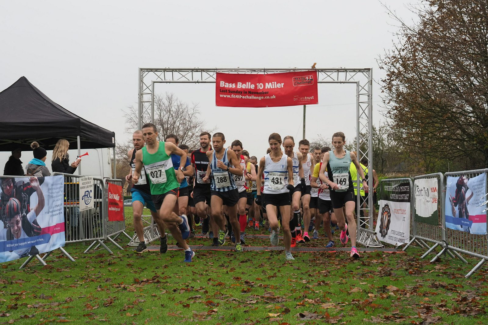 A group of runners start a race, passing under a red banner that reads "Bass Belle to Miller." The ground is grassy, with some autumn leaves scattered around. Spectators and banners line the sides of the path. The sky is overcast.