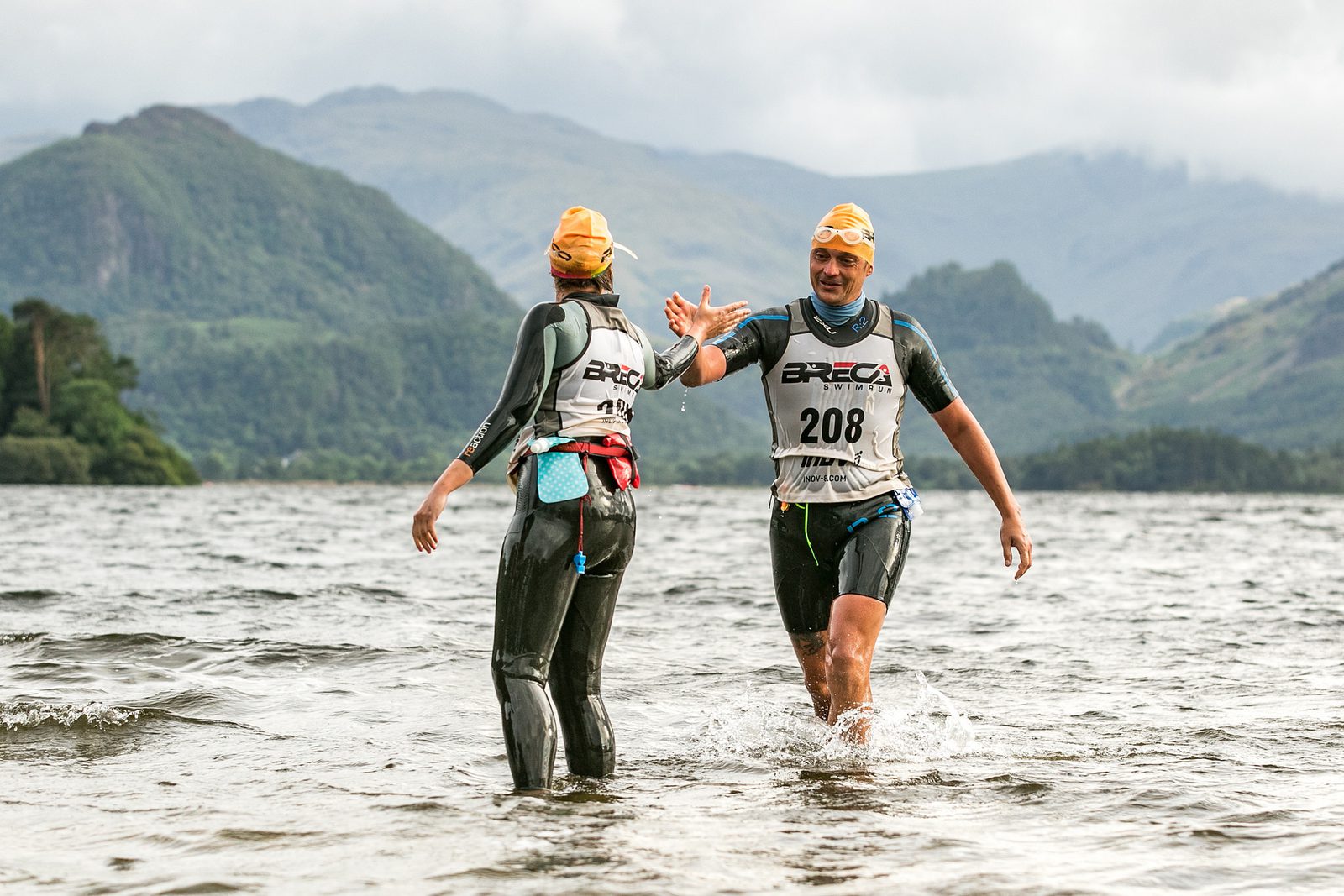 Two athletes wearing swim caps and wetsuits high-five each other while standing in water. They have race numbers on their bibs and appear to be in a mountainous and hilly area with clouds in the sky above. The landscape suggests a challenging outdoor race.