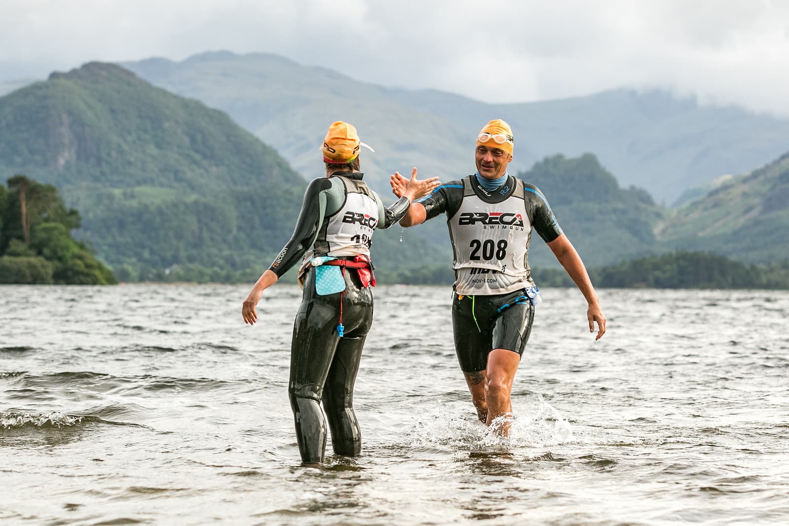 Two triathletes wearing yellow swim caps and numbered white vests high-five each other while standing in a body of water. They are both dressed in wetsuits. The background features lush green mountains under a cloudy sky.