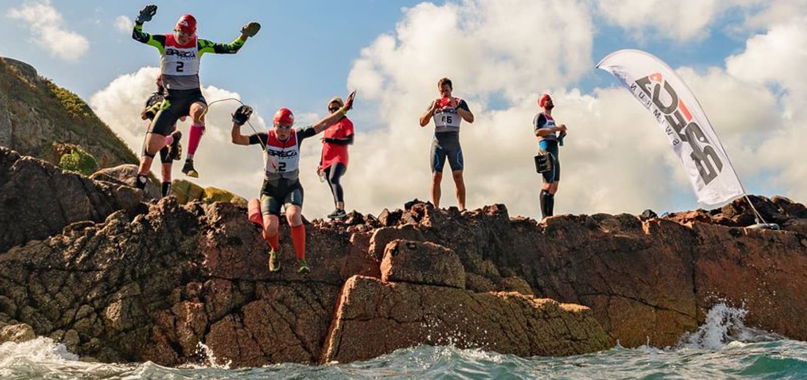 Several athletes wearing numbered jerseys and caps jump off a rocky cliff into the water below, participating in an outdoor sporting event with a blue sky and white clouds in the background. A flag bearing a logo is visible on the right side of the cliff.