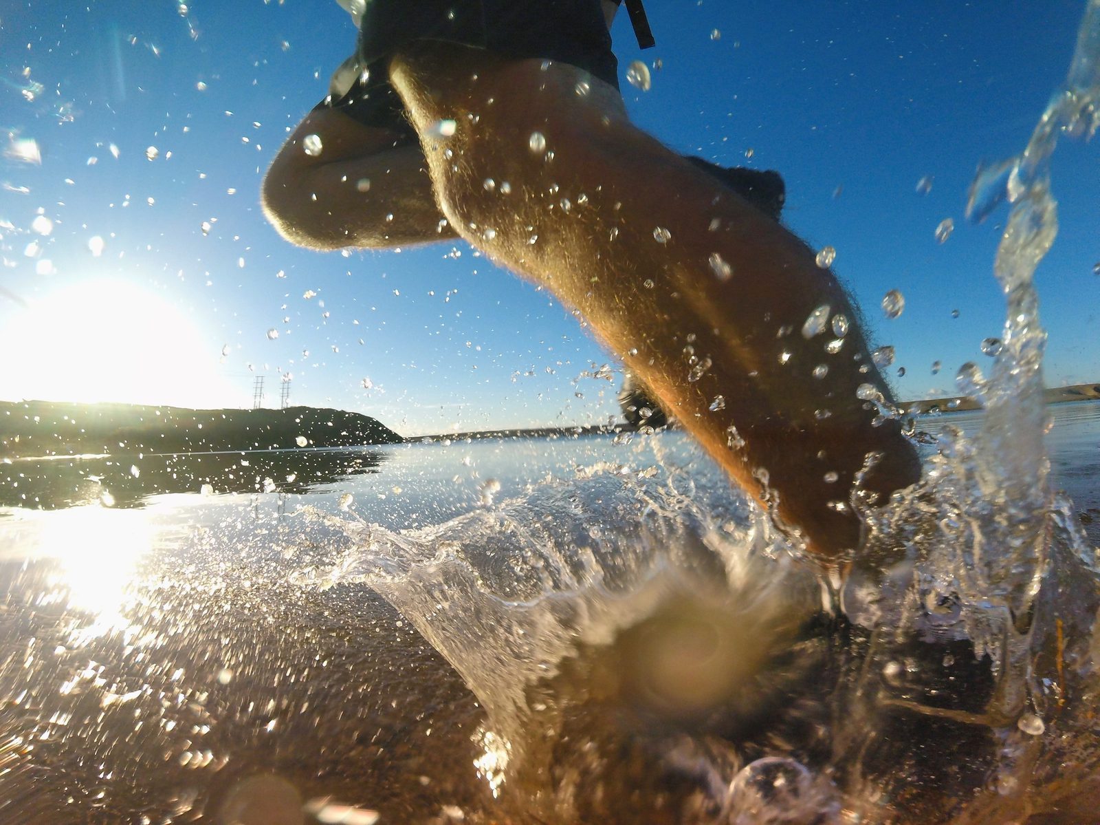 Close-up shot of a person running through shallow water at the beach, with water splashing around their feet. The sun is low on the horizon, casting a bright light and creating a serene, scenic backdrop of the ocean and distant coastline.