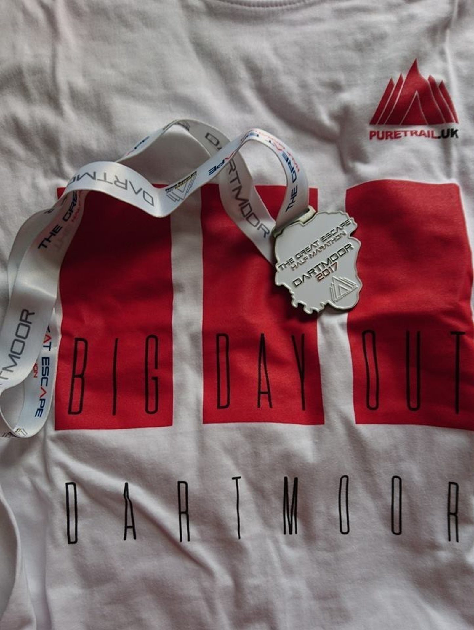 A white t-shirt with "Big Day Out Dartmoor" printed in large red and black letters, along with the logo "PureTrail UK". The shirt also features a medal on a white ribbon with blue text, indicating participation in "The Great Escape Dartmoor 2017".