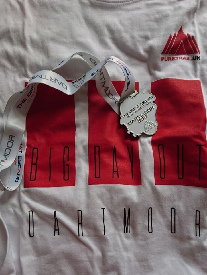 A white t-shirt with "Big Day Out Dartmoor" printed in large red and black letters, along with the logo "PureTrail UK". The shirt also features a medal on a white ribbon with blue text, indicating participation in "The Great Escape Dartmoor 2017".