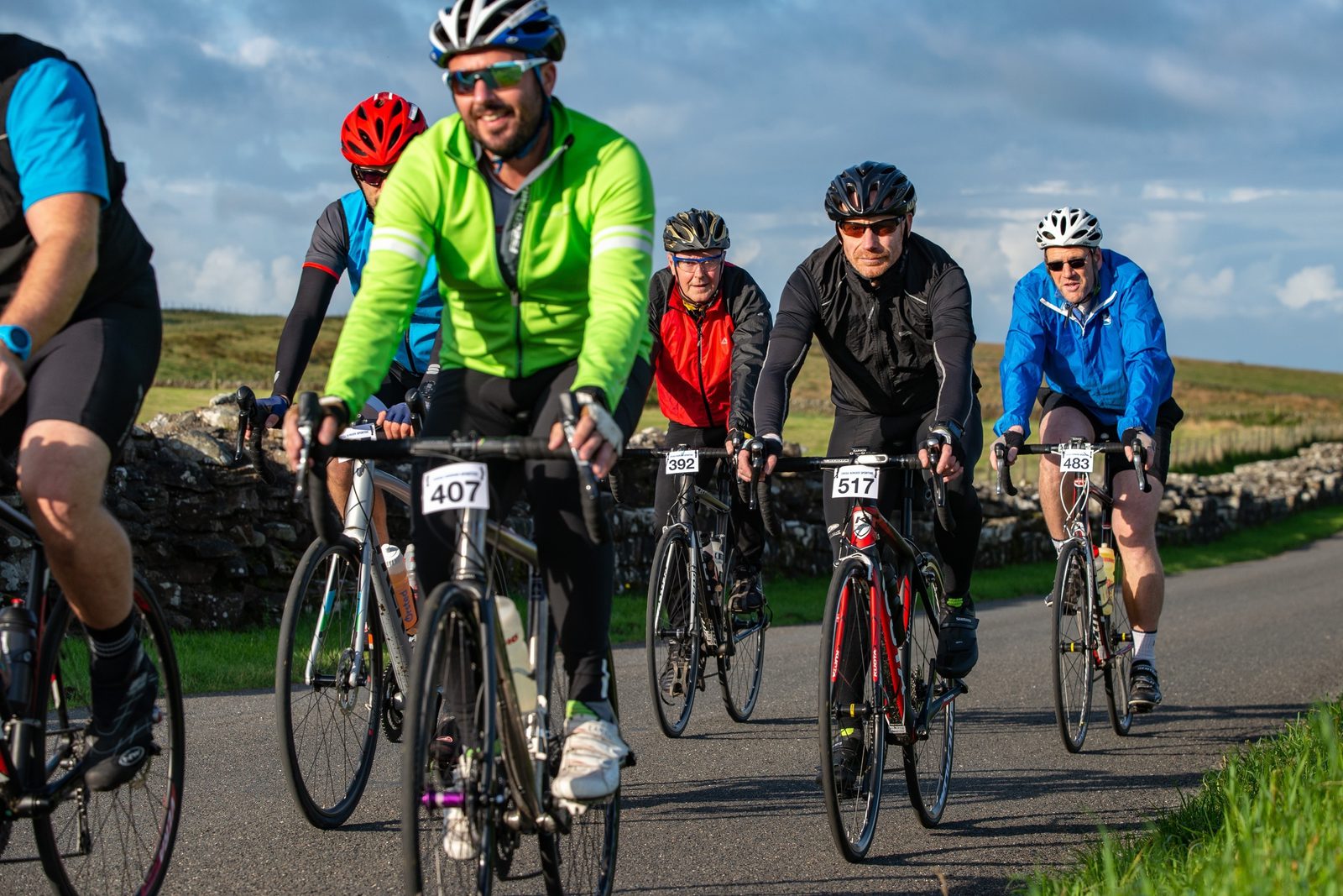A group of cyclists ride on a paved road during a biking event, each wearing helmets and cycling attire. The riders have numbers attached to their bikes, indicating participation in an organized event. A stone wall and green fields are visible in the background under a clear sky.